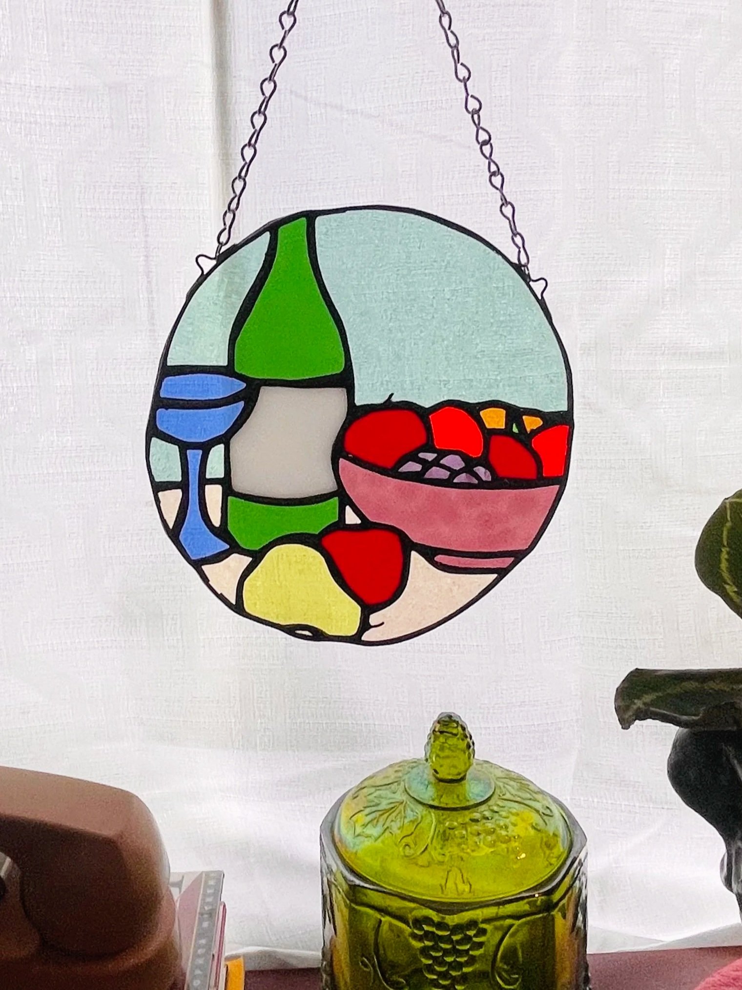 Stained glass hanging panel with various colored sections including green, blue, red, and yellow, attached by two chains, above a yellow glass vase with intricate plant-like designs, on a white surface.