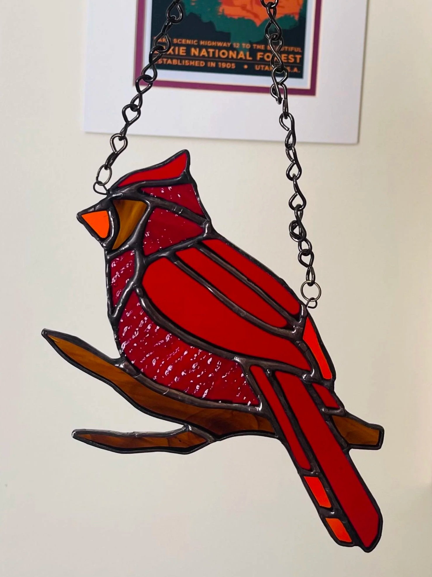 A stained glass art piece of a cardinal bird hanging on a chain, with a scenic highway poster in the background.