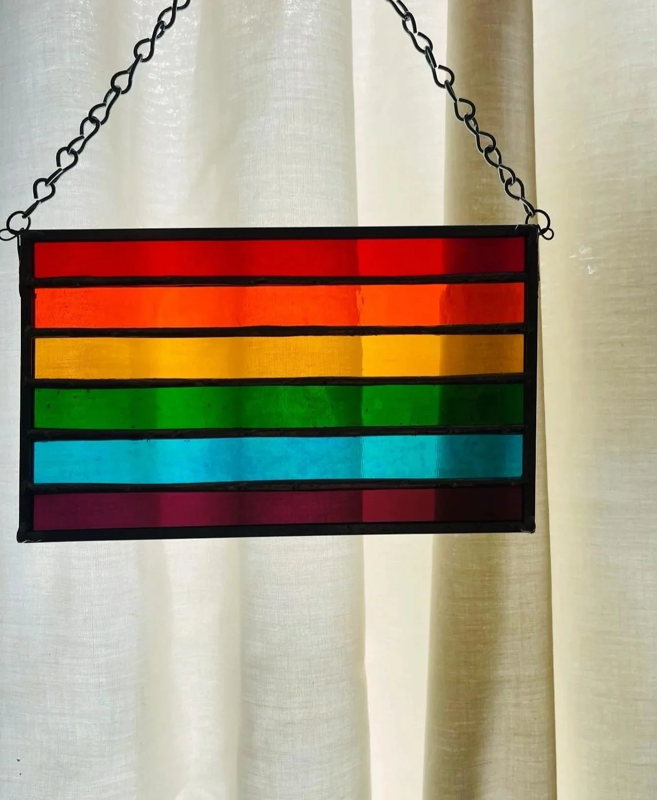 Colorful stained glass panel with horizontal stripes in red, orange, yellow, green, blue, and purple to depict a gay pride flag, hanging from a chain against a light-colored curtain background.