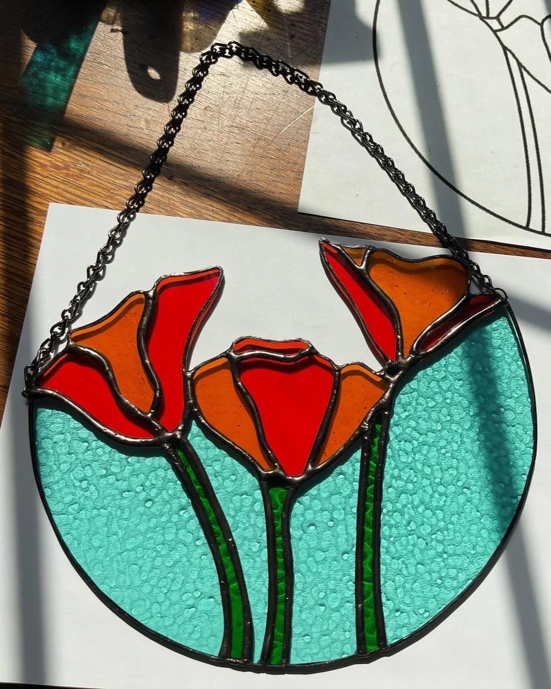 Colorful stained glass artwork featuring three orange and red flowers with green stems on a circular blue background.