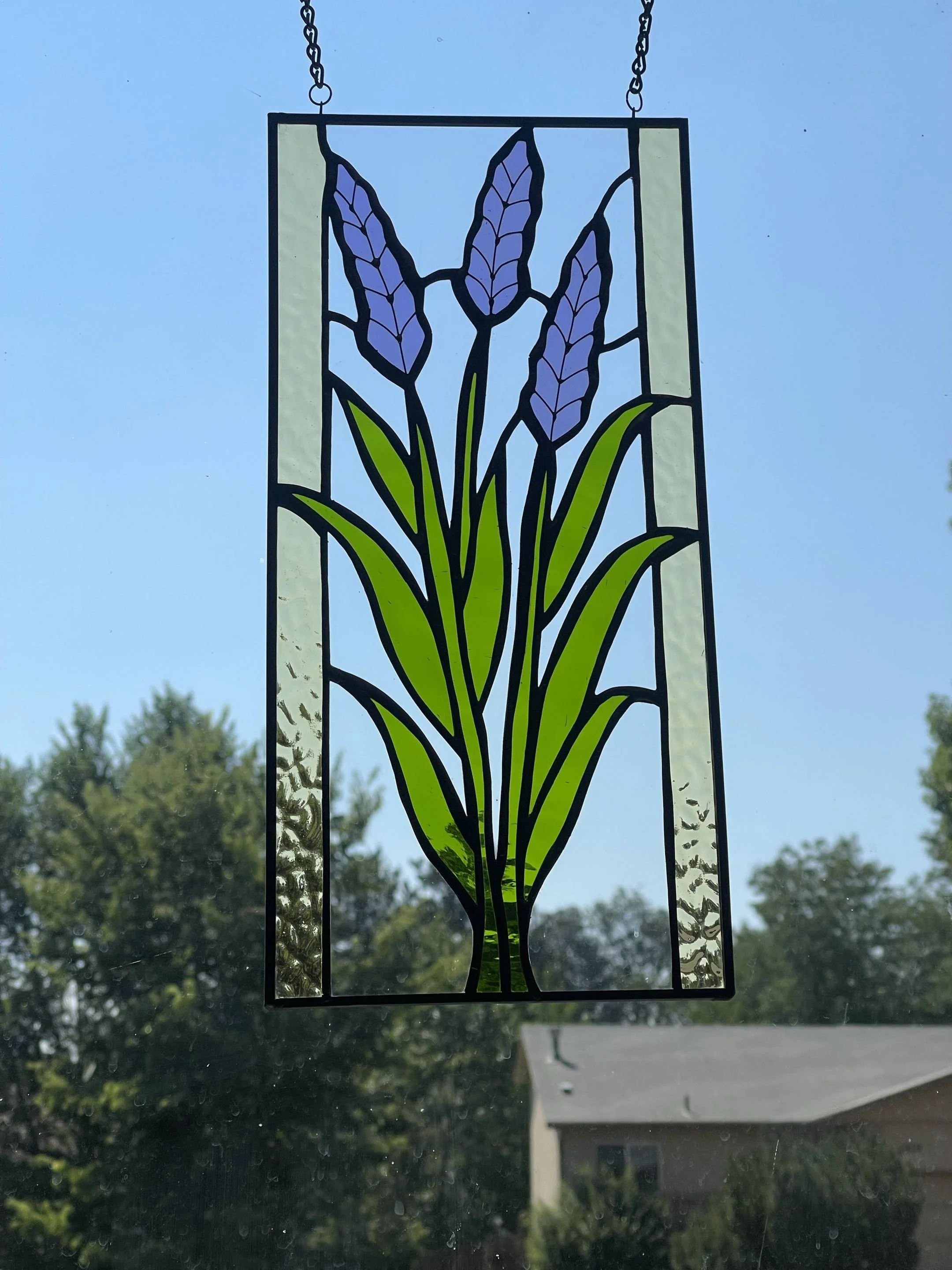 Stained glass window art of lavender with green leaves, hanging against a blue sky with trees and a house in the background.