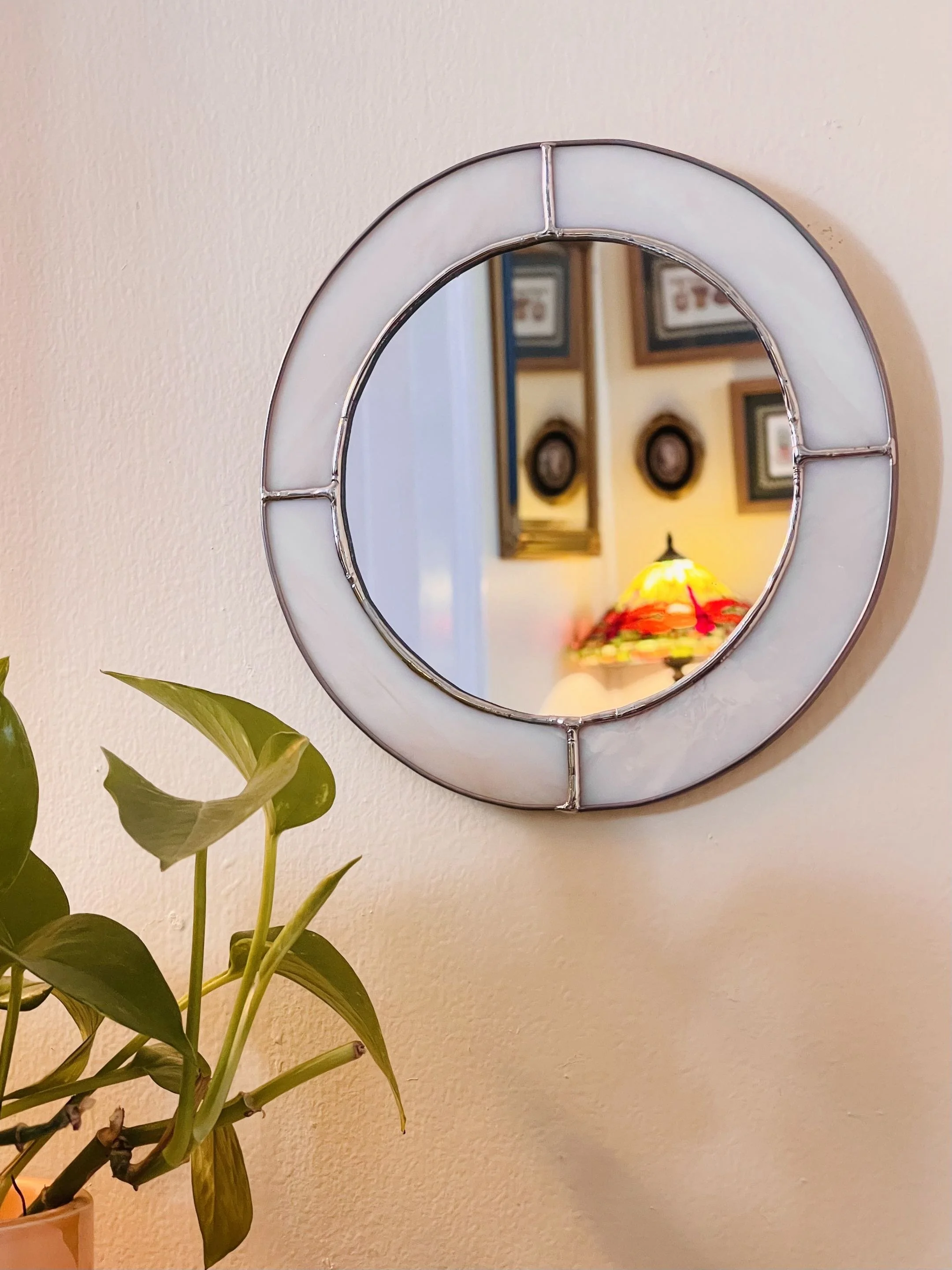 A round mirror mounted on a beige wall, reflecting a room with framed pictures on the wall and a colorful lampshade on a table. A green leafy plant is in the foreground to the left.