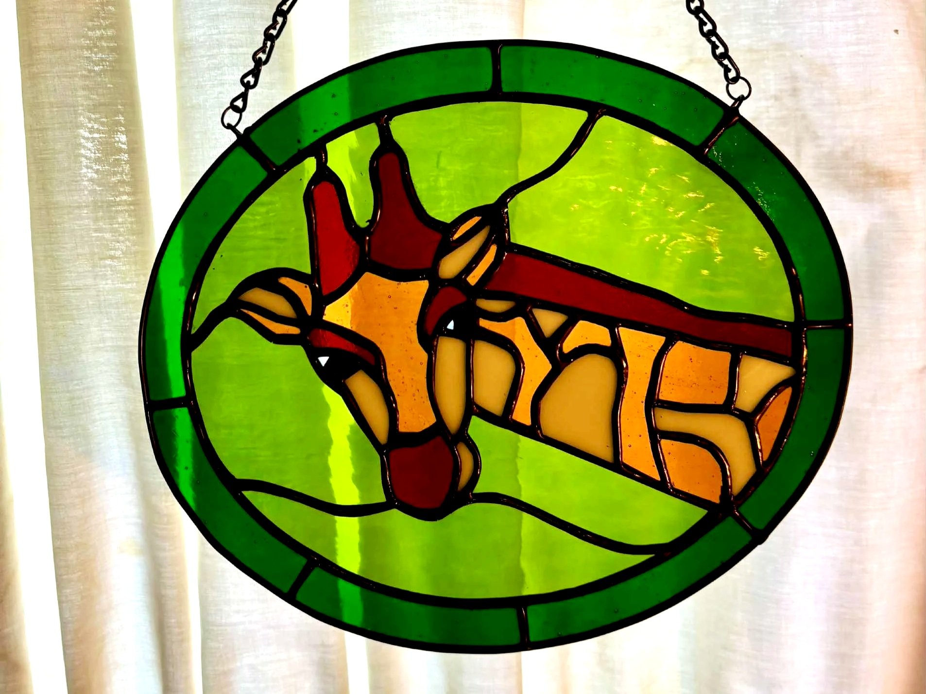 Stained glass artwork of giraffe with hues of green in oval setting.
