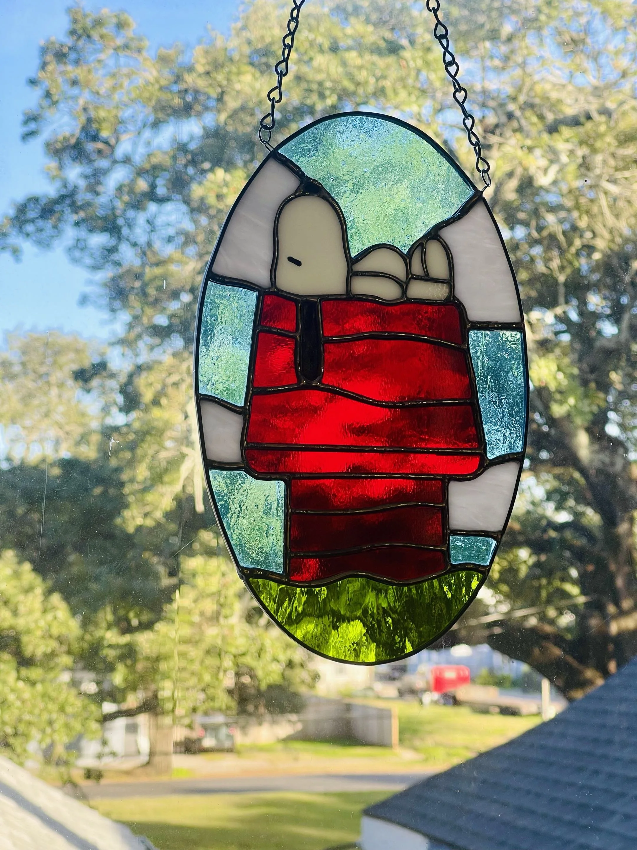 A stained glass ornament hanging in a window, depicting a snoopy the dog laying atop his red doghouse staring up at a blue sky. 