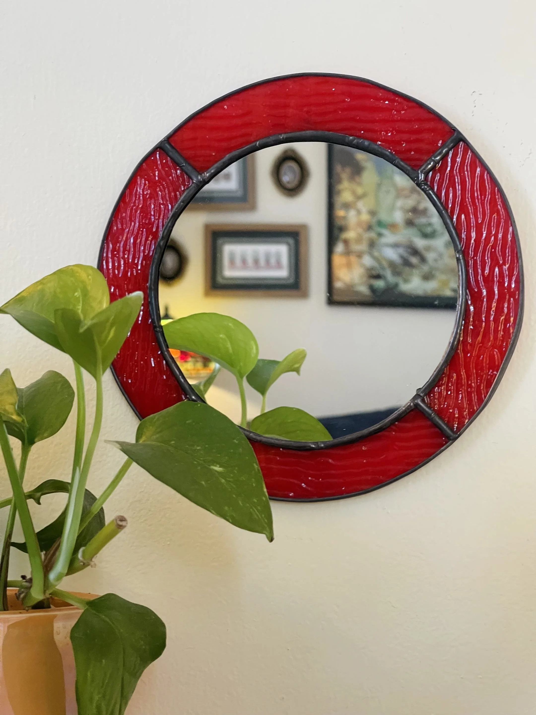 Red stained glass mirror with black metal frame hanging on a beige wall, partially reflecting framed pictures and a clock. Green leafy plant in a yellow pot is in the foreground.