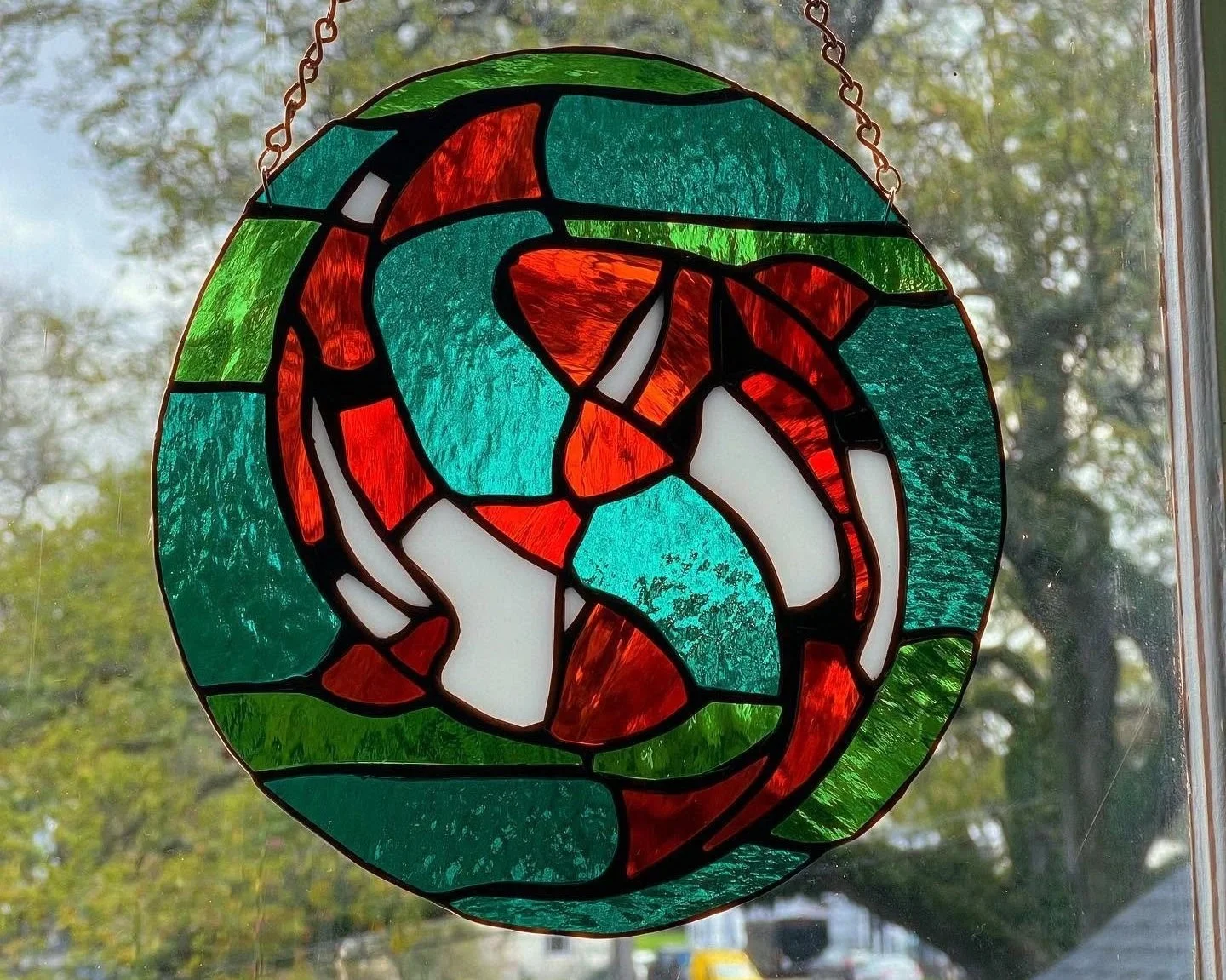 Colorful round stained glass ornament with two koi fish circling each other hanging in front of a window with trees outside.