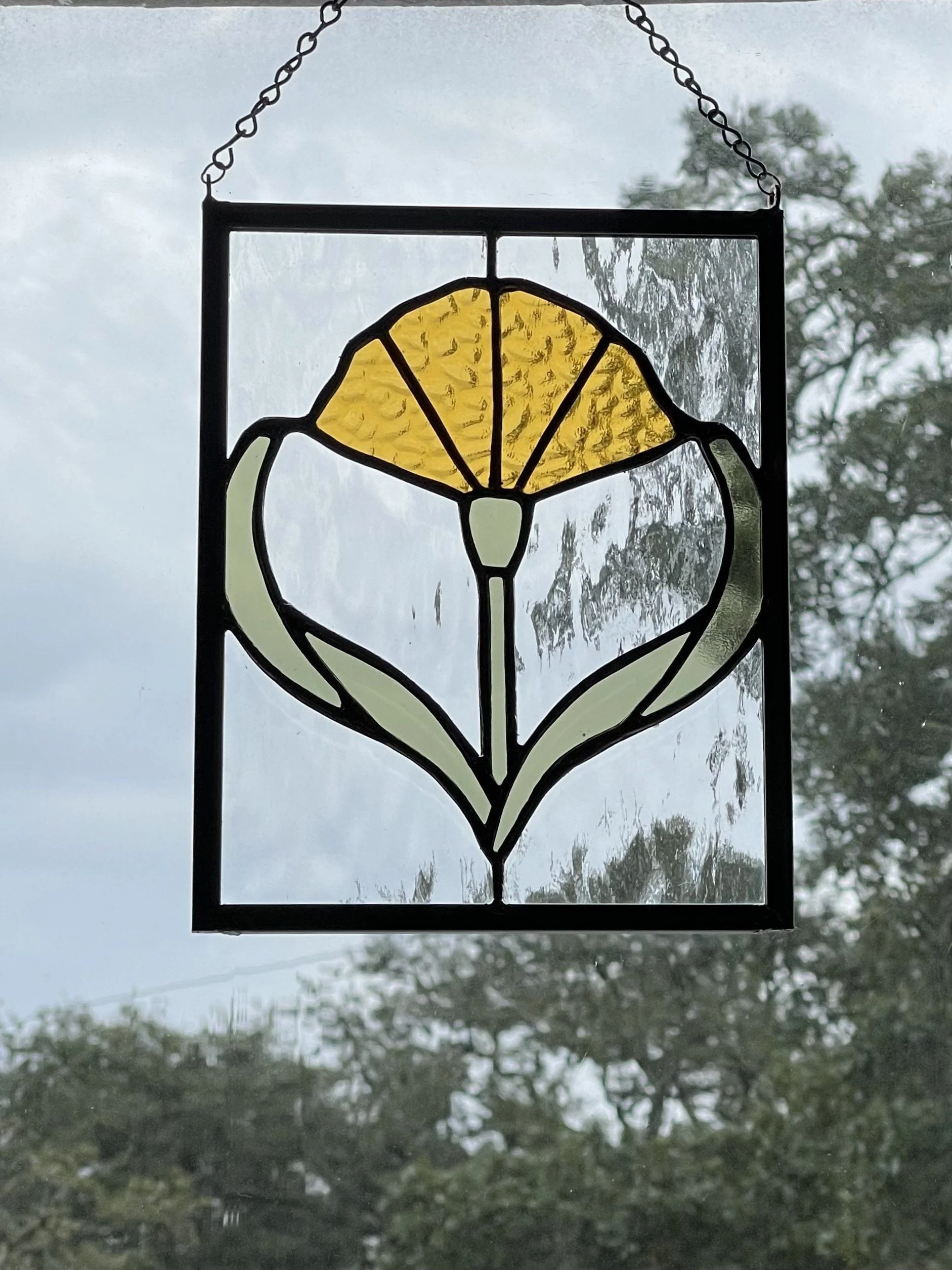 Stained glass hanging decoration featuring a yellow flower design, suspended by a chain, with a cloudy sky and trees in the background.