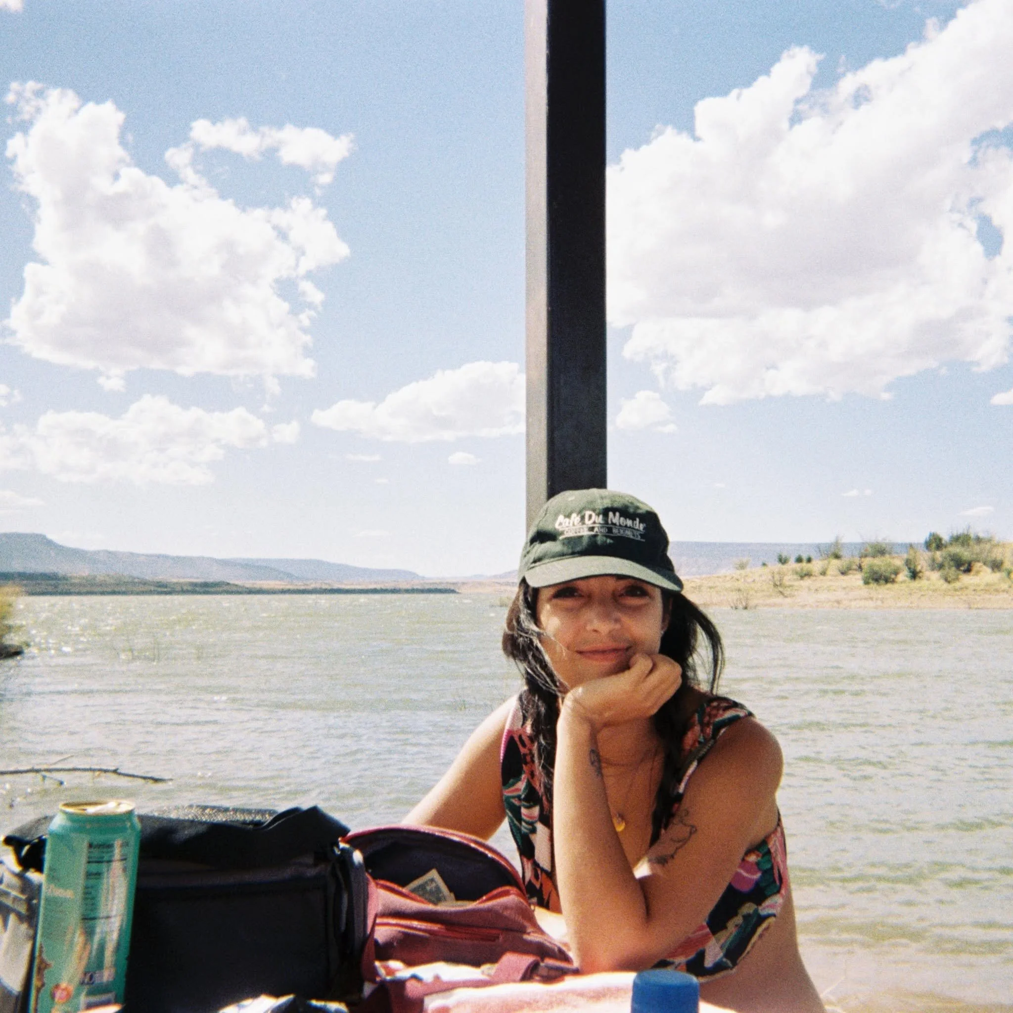 A woman with dark hair, wearing a colorful floral top and a baseball cap, sitting outdoors at a beach with mountains and a body of water in the background. There are some cans and a bag or backpack near her.