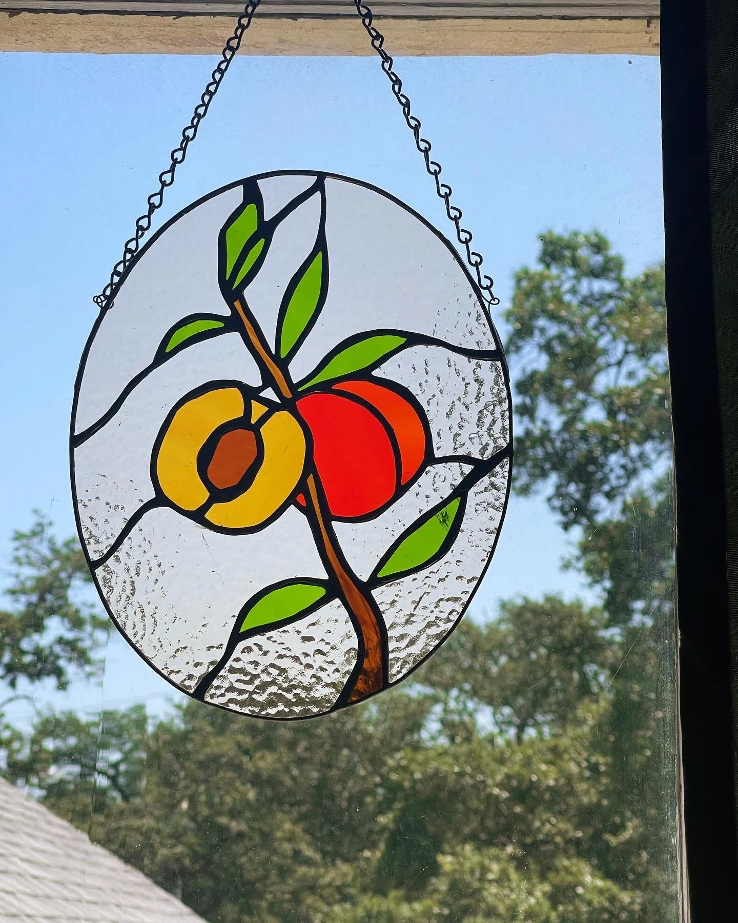 Stained glass artwork with a peach, green leaves, and a brown stem, hanging by a chain, against a blue sky and trees in the background.