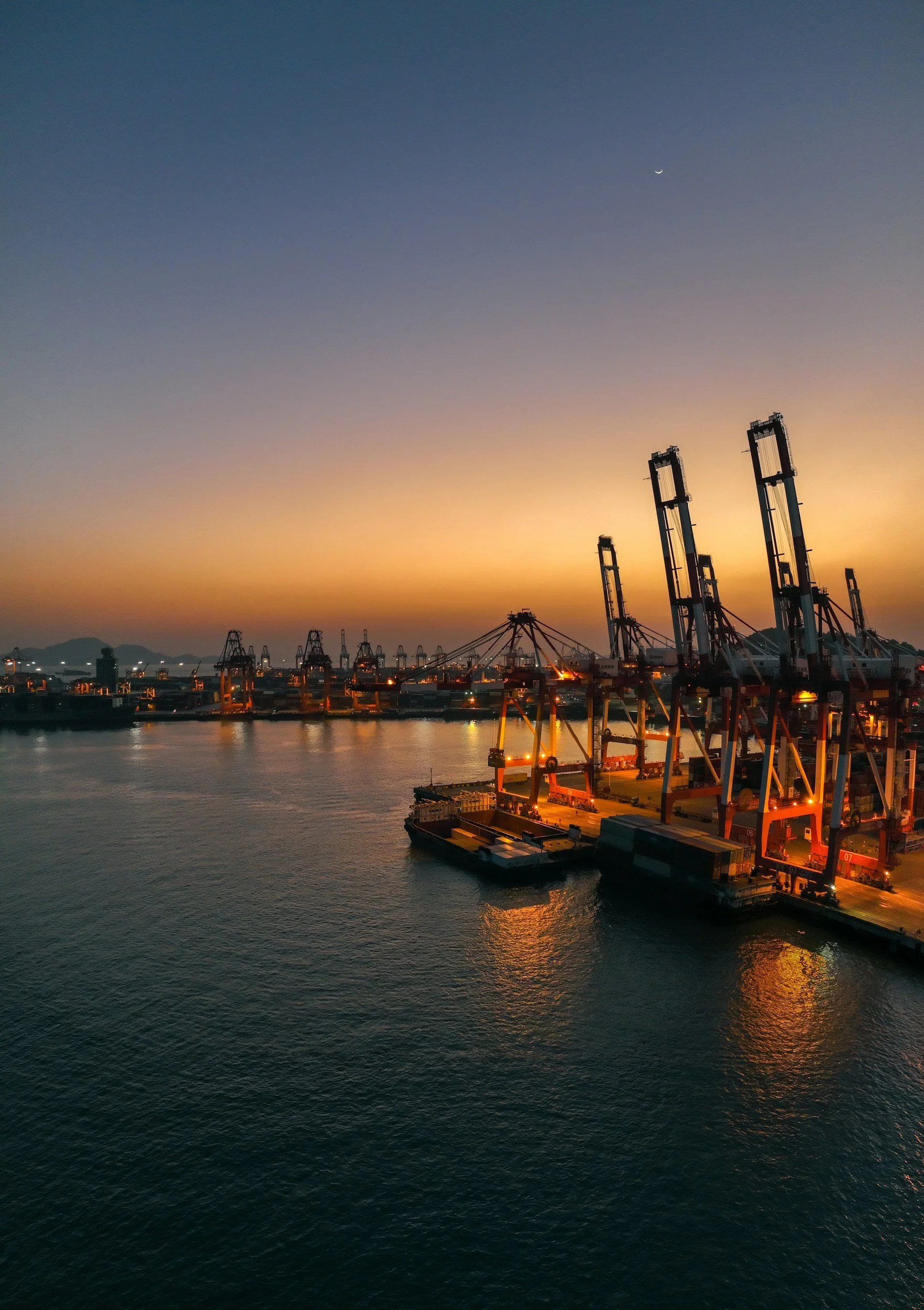 Several large cargo cranes at a port during sunset, with the sky transitioning from orange to blue and a crescent moon visible, and calm water reflecting the scene.