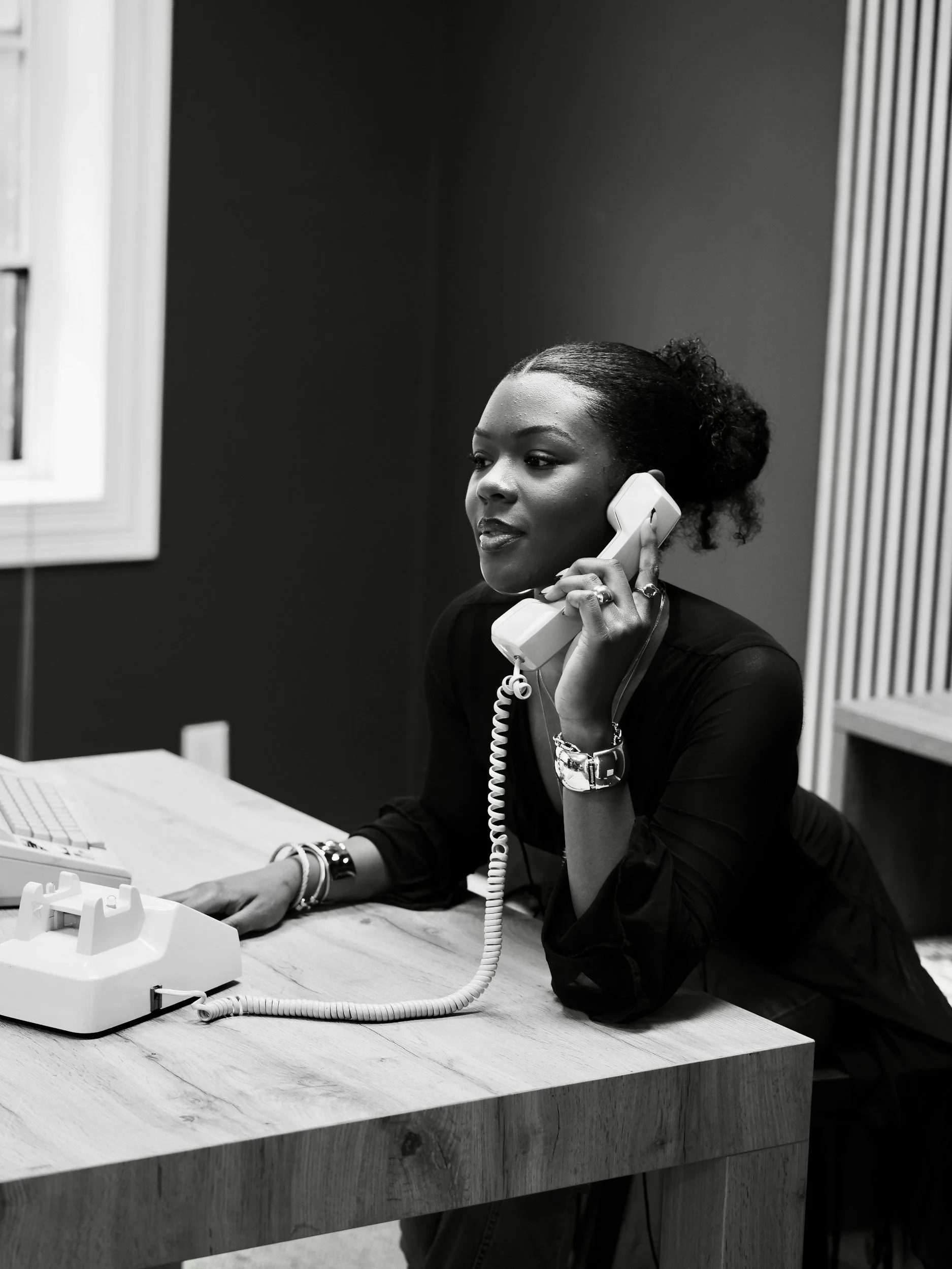 A black-and-white photo of a woman with dark skin and curly hair in a bun, sitting at a desk, speaking on a landline phone, with a focused expression. She is wearing a dark top and accessorized with bracelets and rings. The desk has a vintage landline phone and a computer keyboard, with a window and patterned wall in the background.