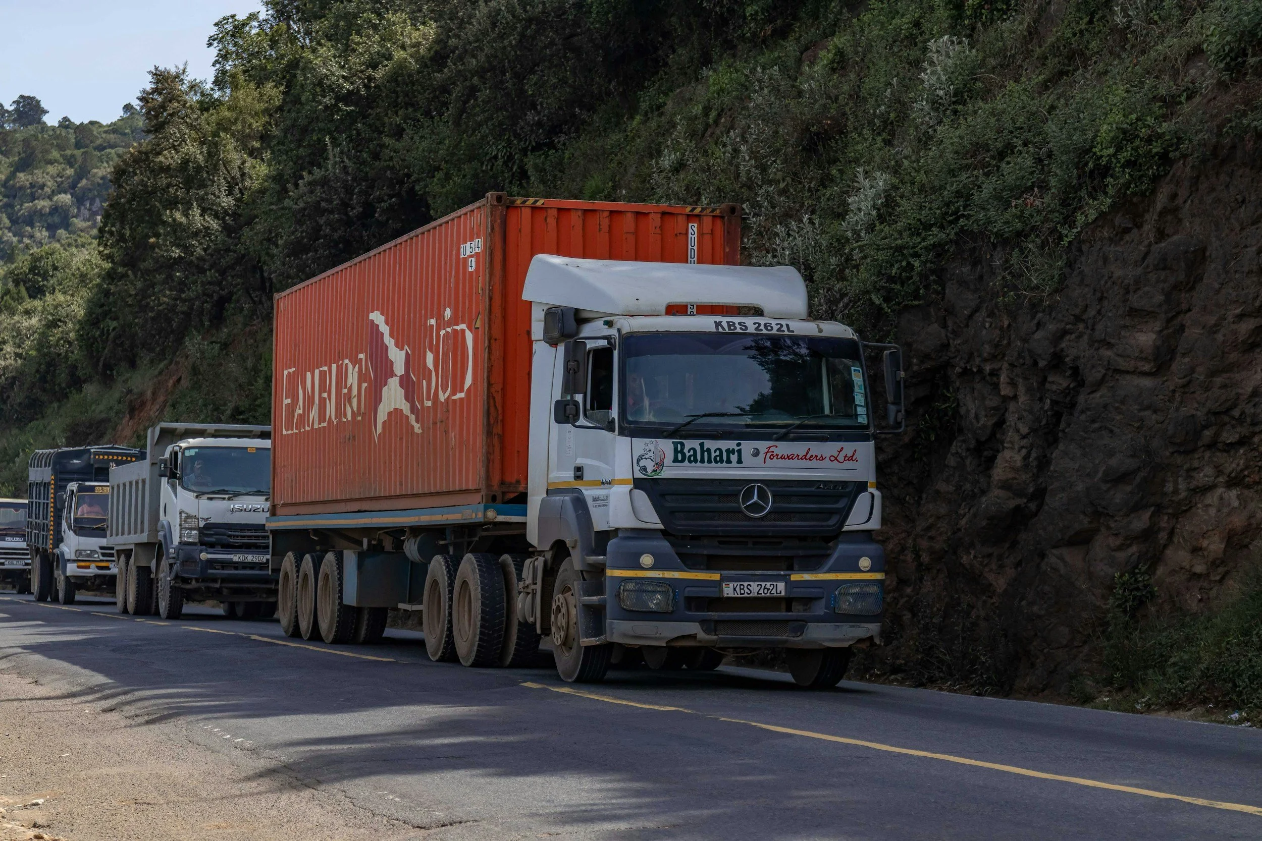 Line of trucks driving on a highway next to a hillside covered with green trees.