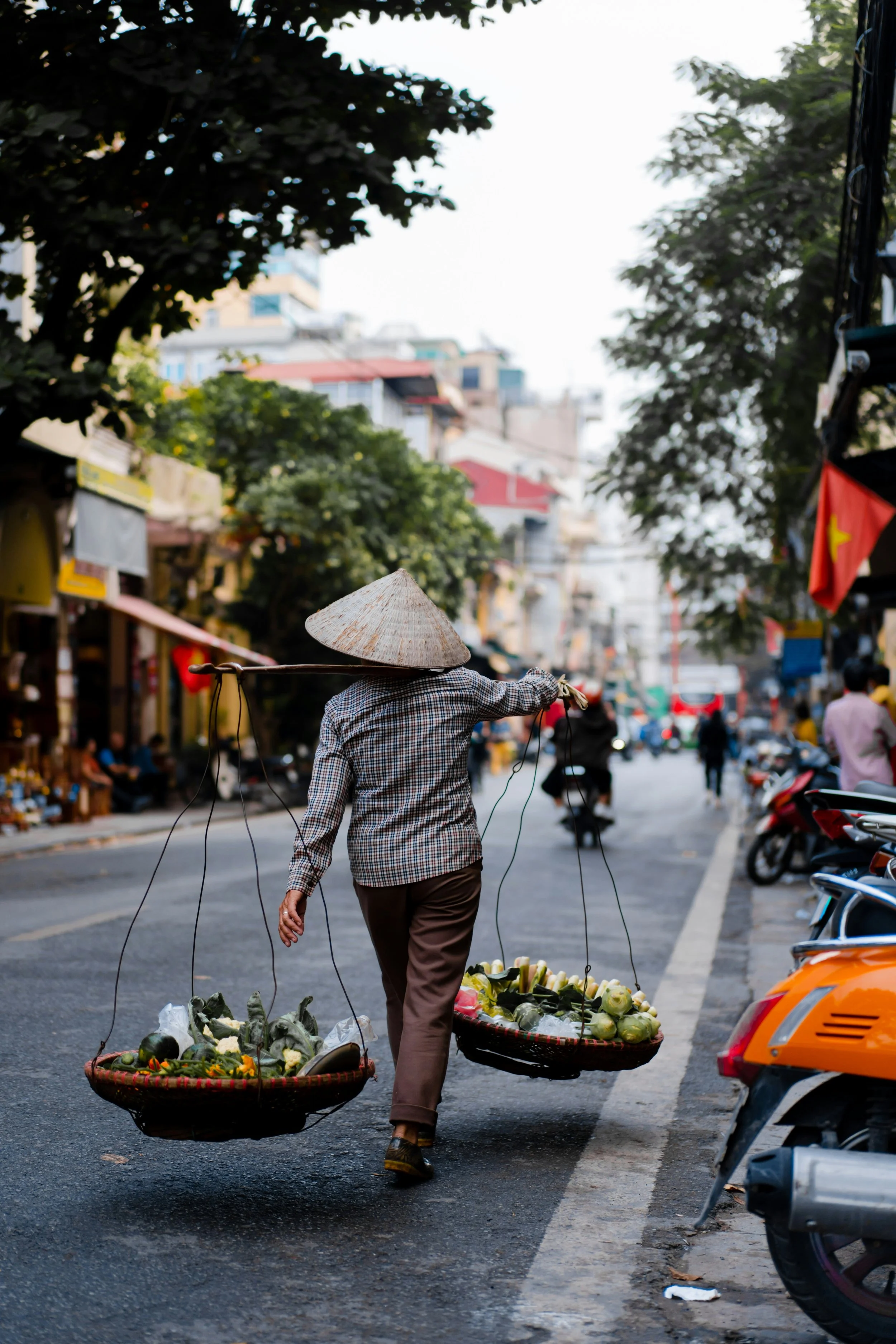 A street scene with a vendor wearing traditional Asian country attire and a conical hat, walking away while carrying baskets of fruits and vegetables. The street is lined with shops, motorbikes, and trees, with a bright sky overhead.