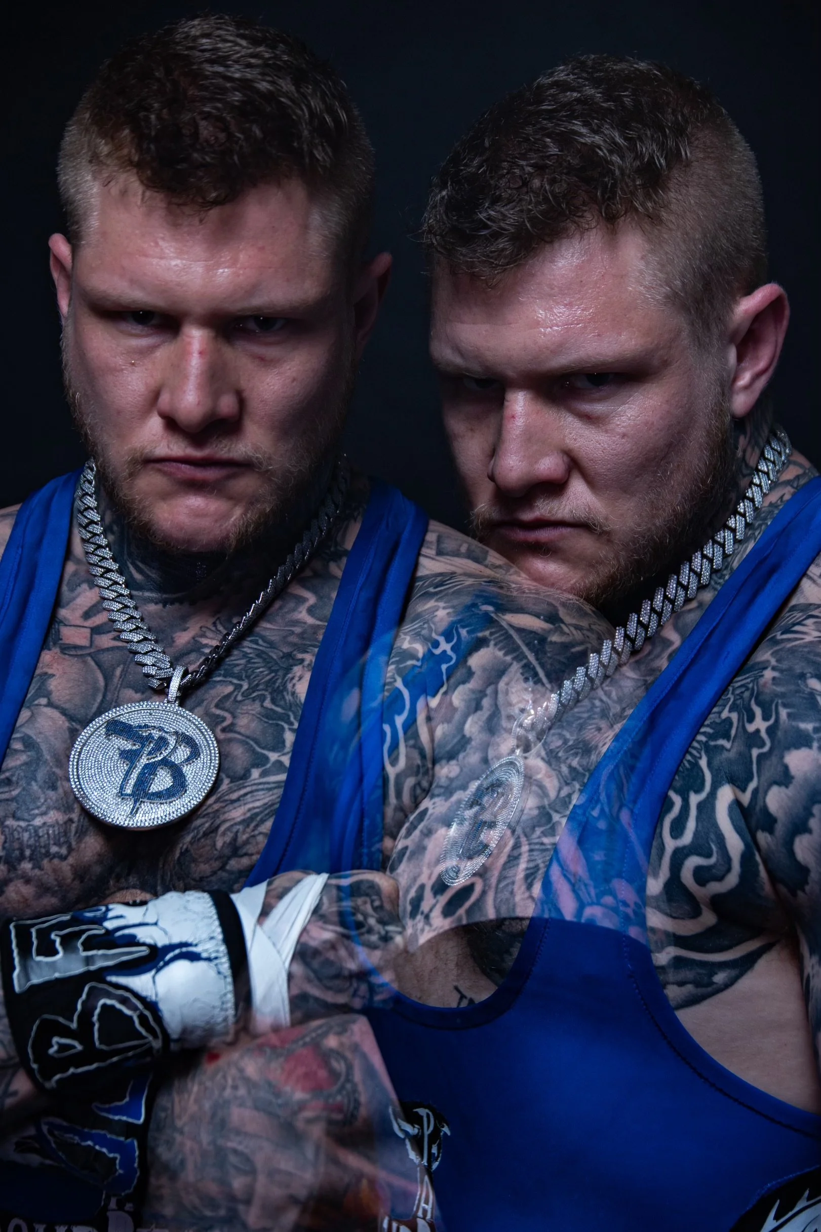 Two muscular men with tattoos wearing blue boxing robes, jewelry, and gloves, posing serious in a dark setting.