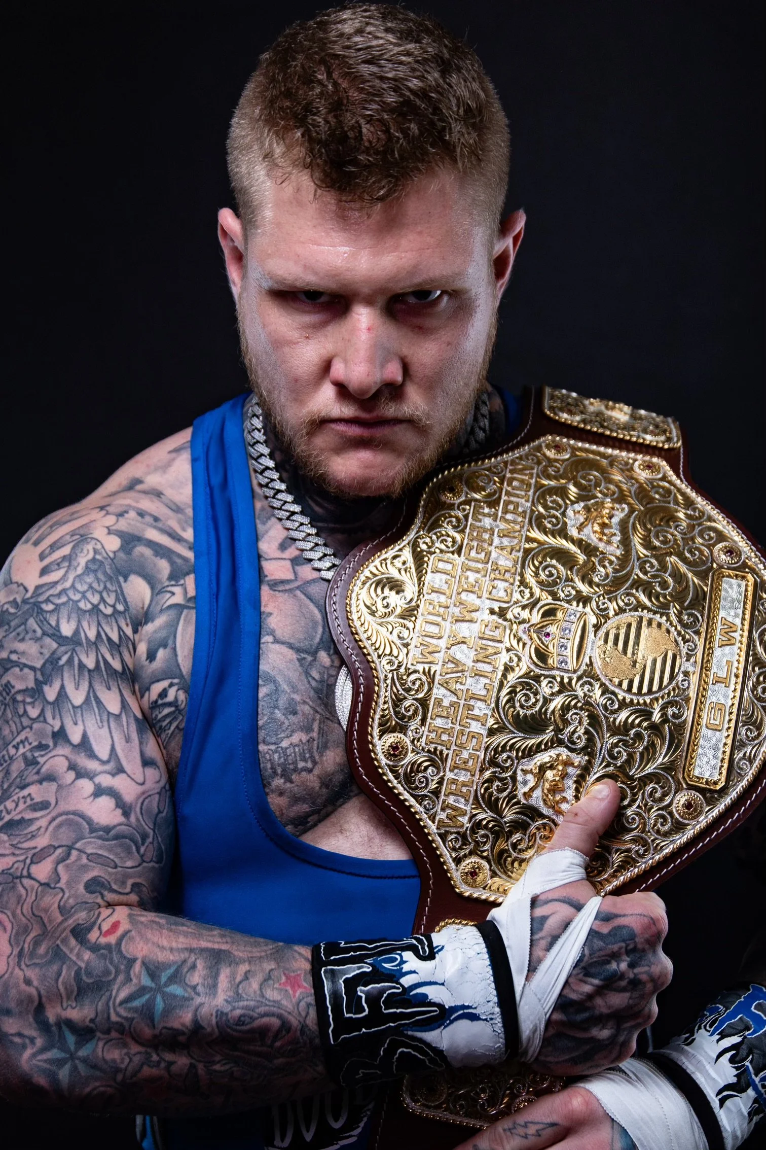 A man with tattoos and a serious expression, wearing a blue tank top and boxing gloves, holding a large ornate championship belt with gold detailing against a dark background.