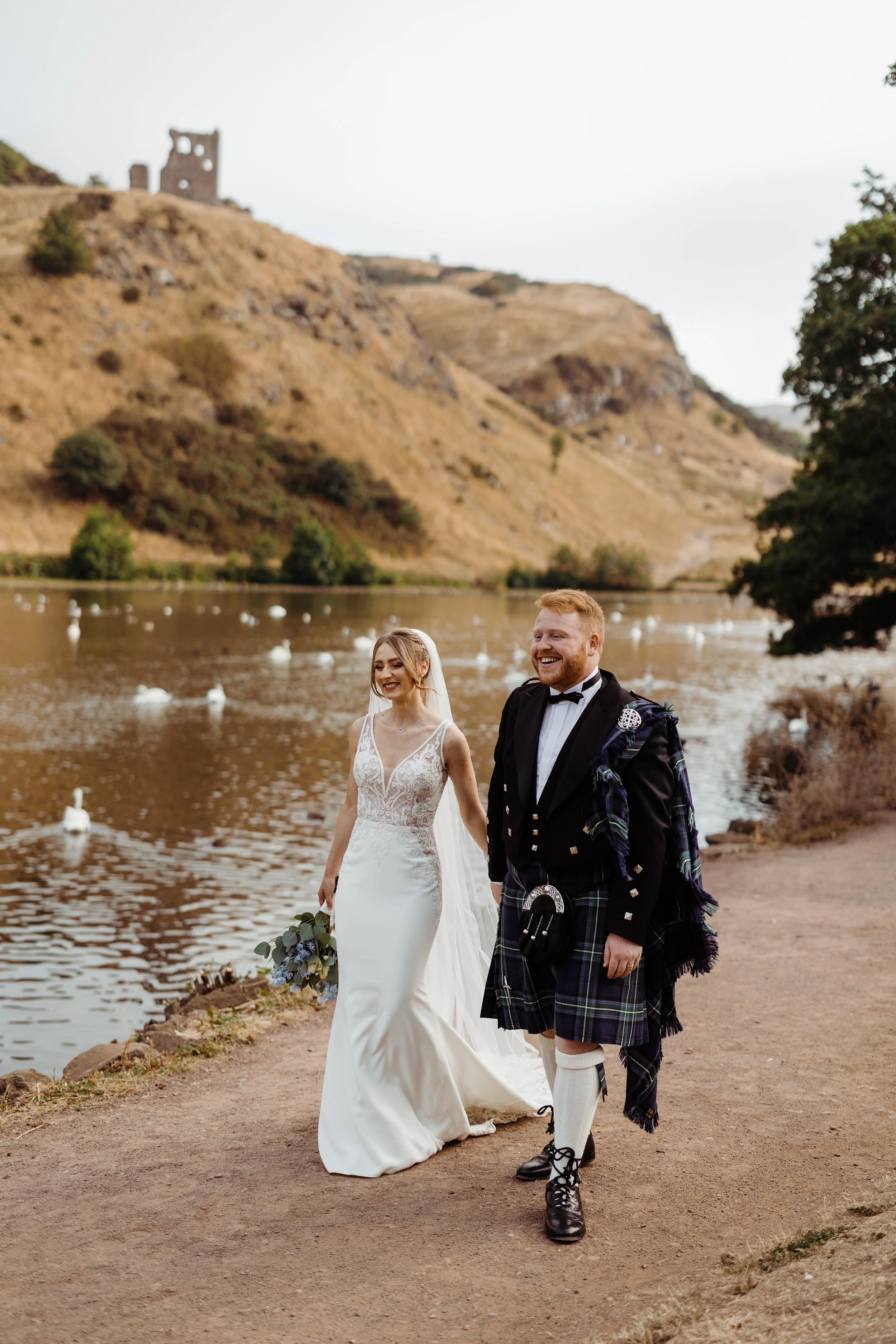A bride and groom walking along a riverbank, smiling. The bride is wearing a white wedding gown and holding a bouquet, and the groom is dressed in traditional Scottish kilt and jacket. The background features a river, white swans, and rugged hills with a historical ruin on top.