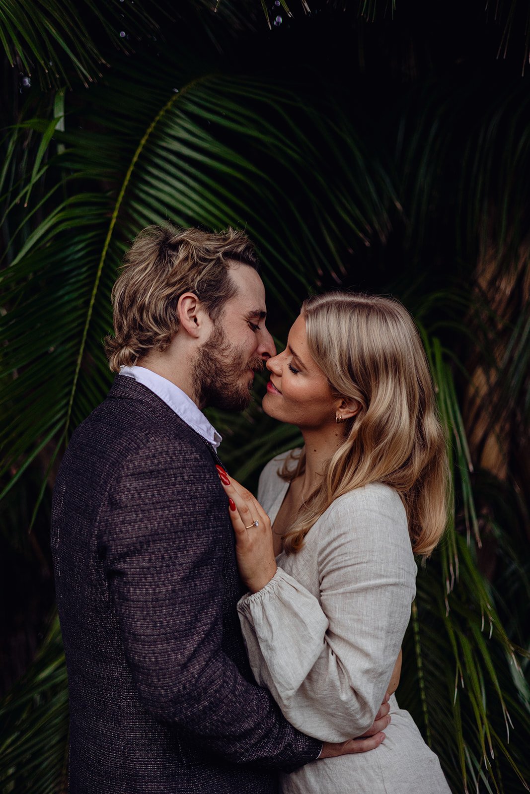 A couple standing close together, smiling and about to kiss, with large green tropical leaves in the background.