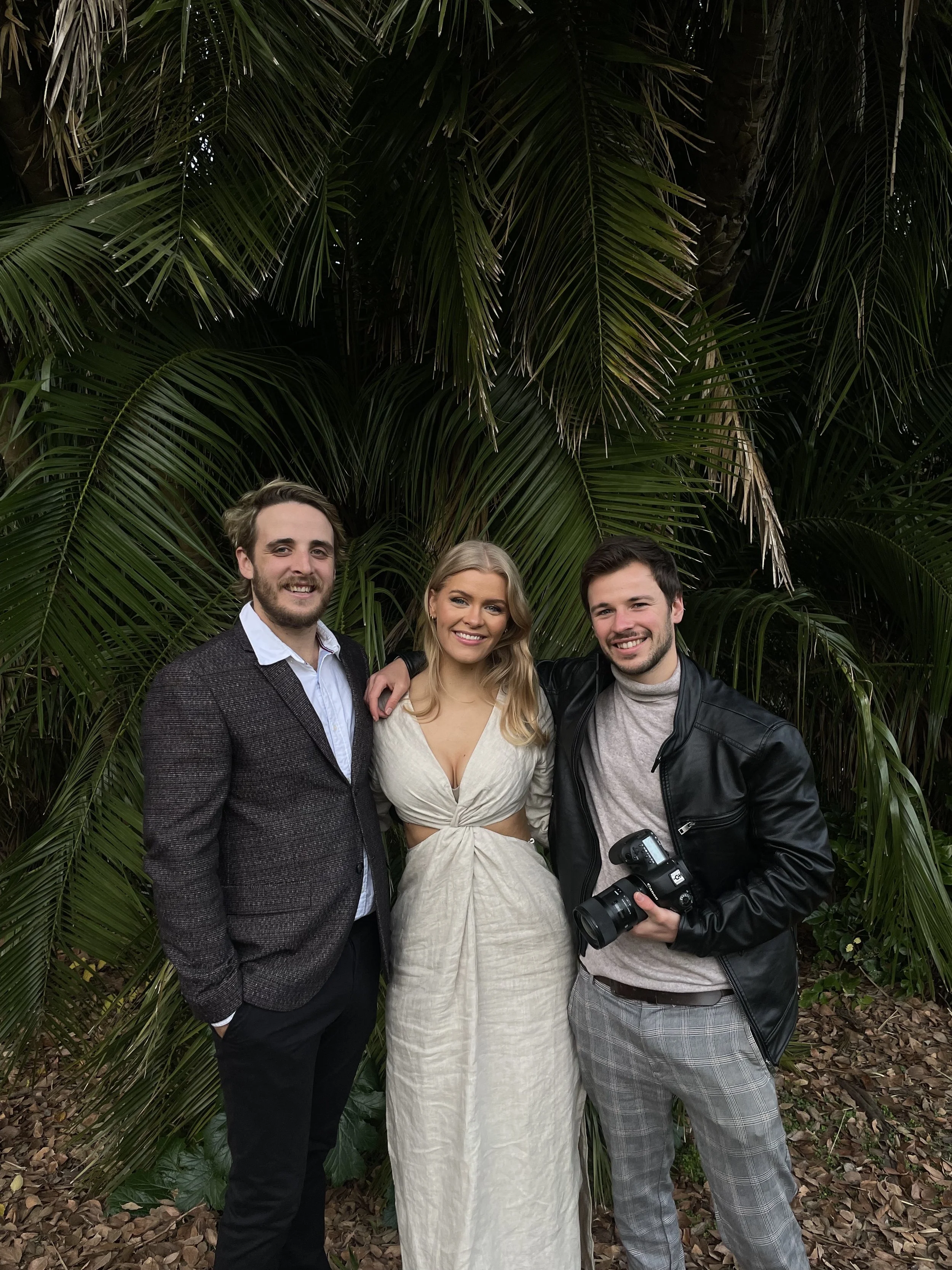 Three young adults, two men and one woman, standing close together outdoors in front of lush green palm trees, smiling at the camera. The man on the left has light brown hair and a beard, wearing a dark blazer over a light shirt. The woman in the middle has long blonde hair, wearing a cream-colored dress. The man on the right has dark hair, wearing a black leather jacket and holding a camera.