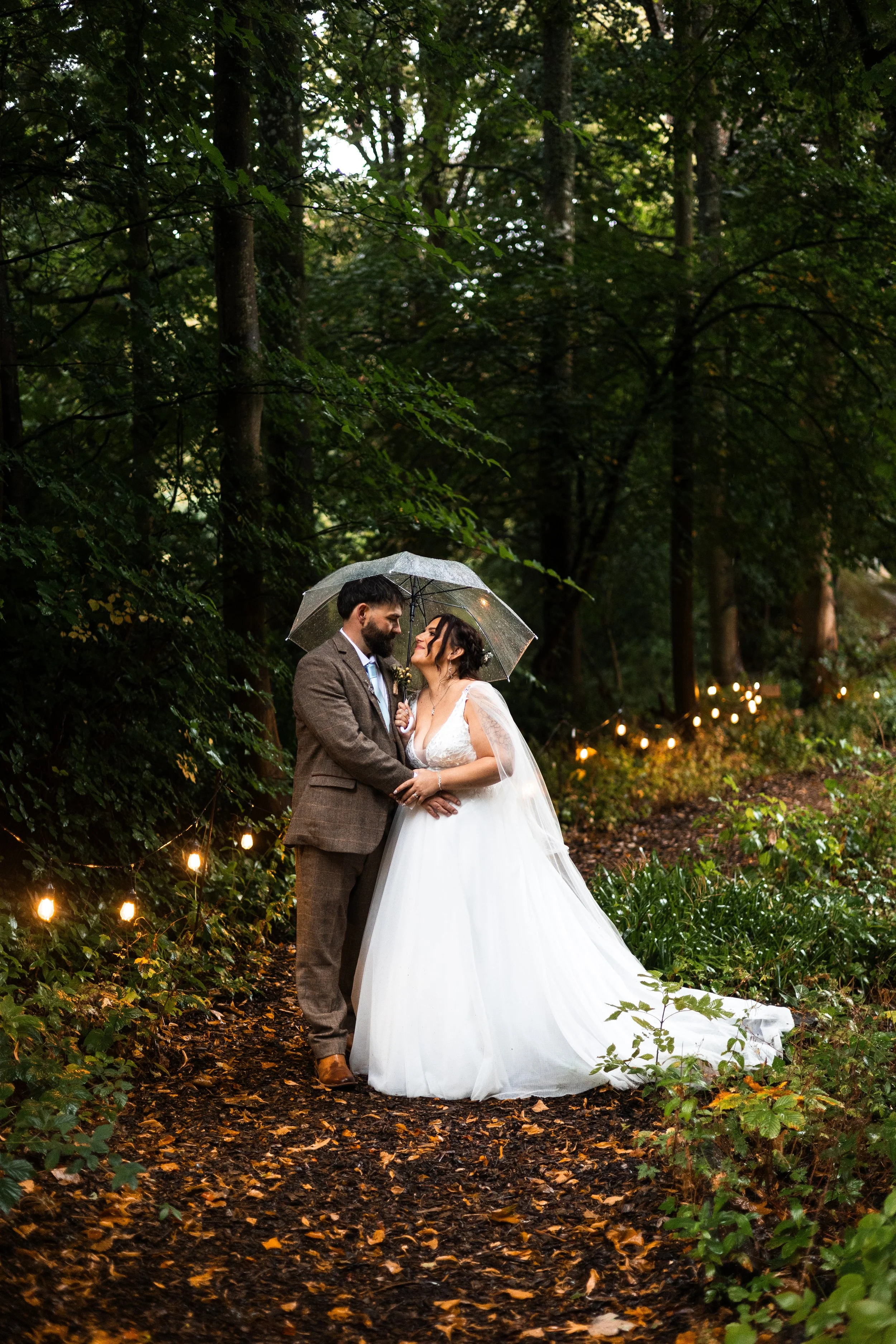 A bride and groom standing close together under a transparent umbrella in an outdoor wooded setting, with string lights hanging in the background, during a rainy evening.