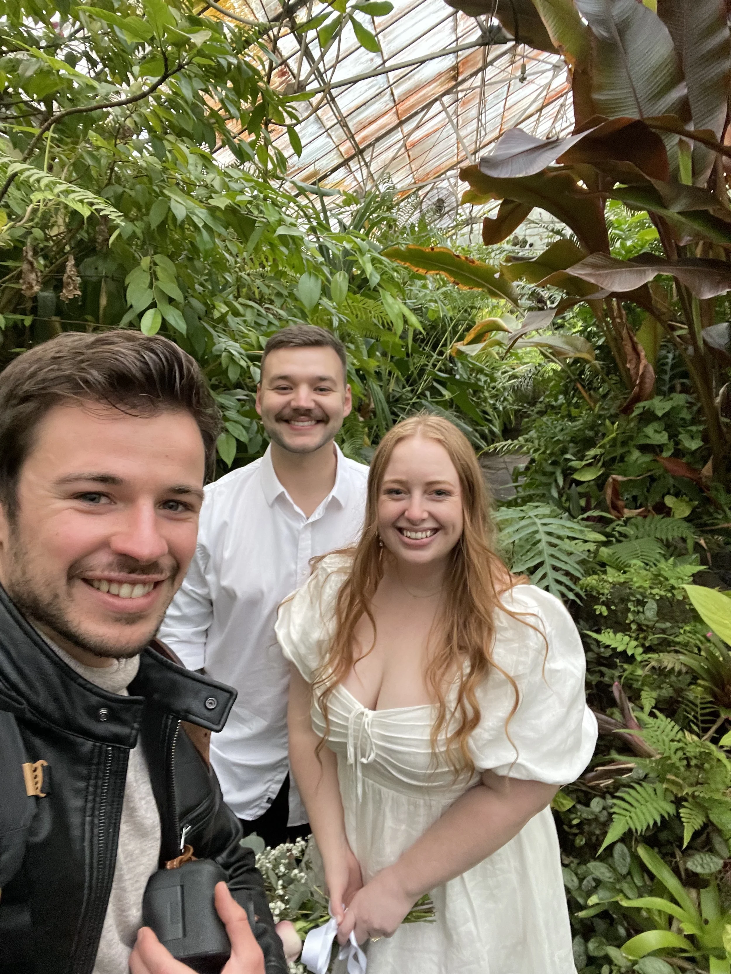 Four people smiling for a photo inside a lush greenhouse. A man on the left holds a camera, with three others standing close together, two women and one man, among dense tropical plants with large green leaves.