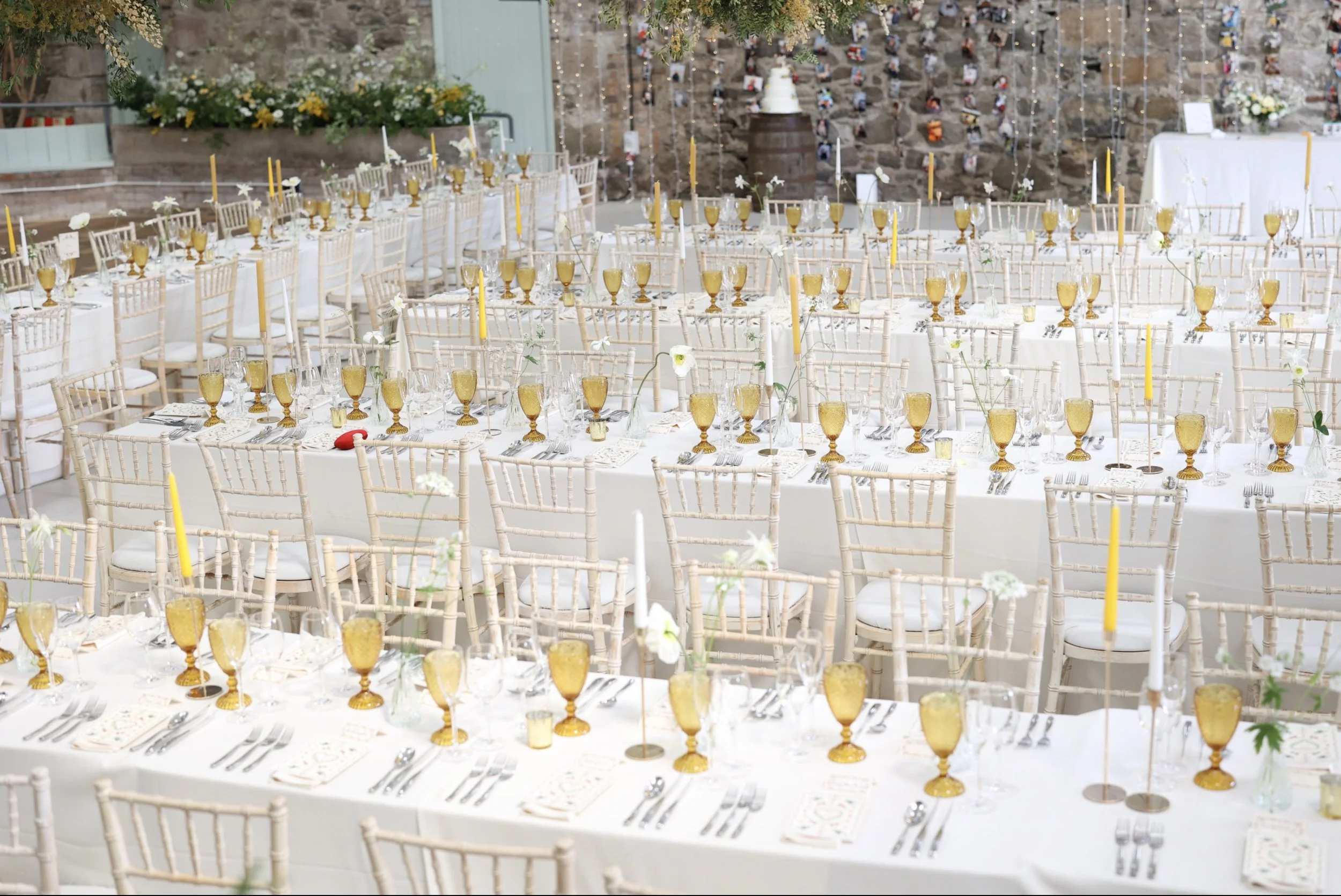 A large outdoor banquet table decorated with white tablecloths, gold goblets, candles, and flowers, set up for a formal event against a stone wall with hanging string lights.