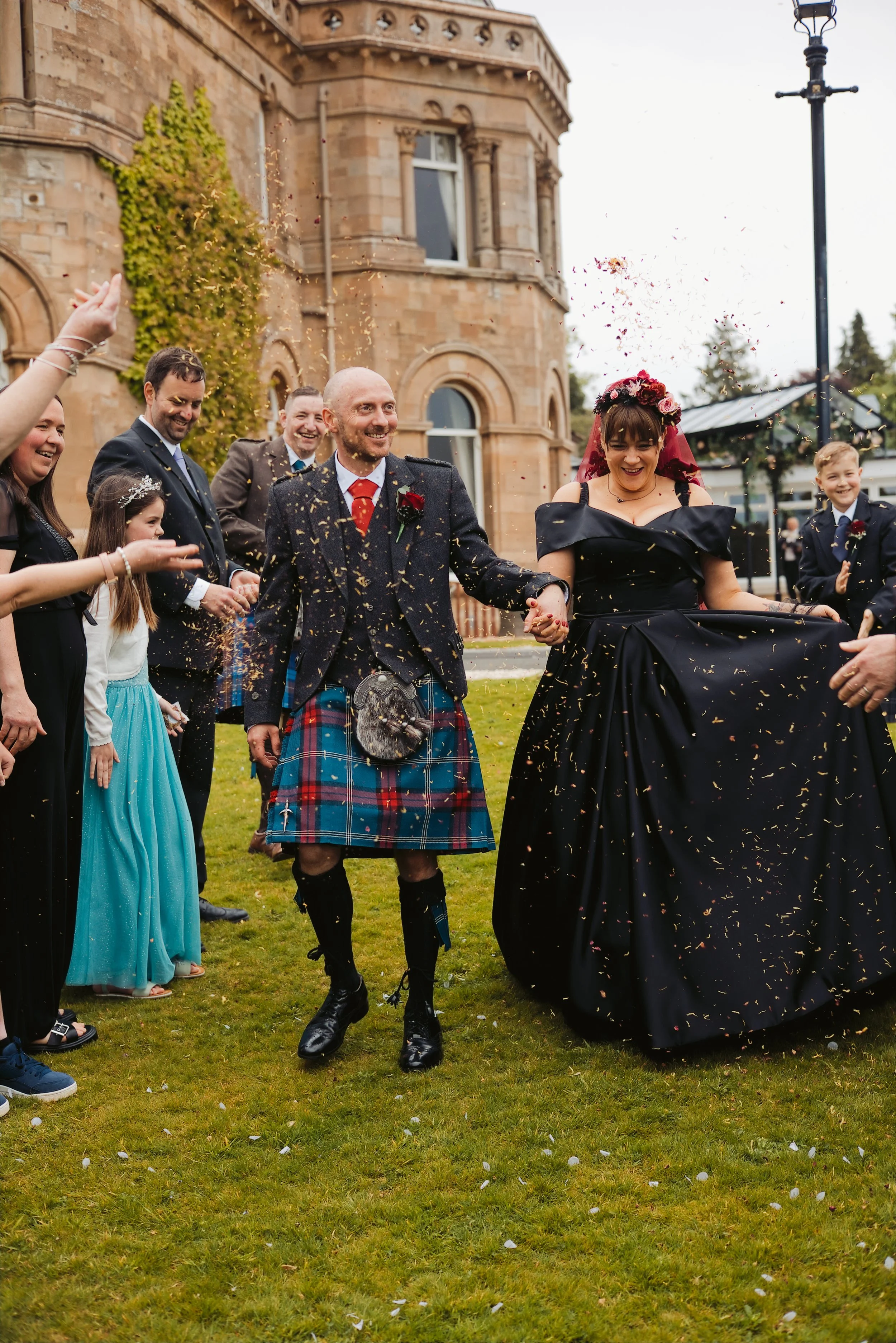 A wedding celebration outdoors with guests throwing confetti, featuring a woman in a black dress with a floral headpiece and a man in Scottish kilt attire holding hands, in front of a historic building.