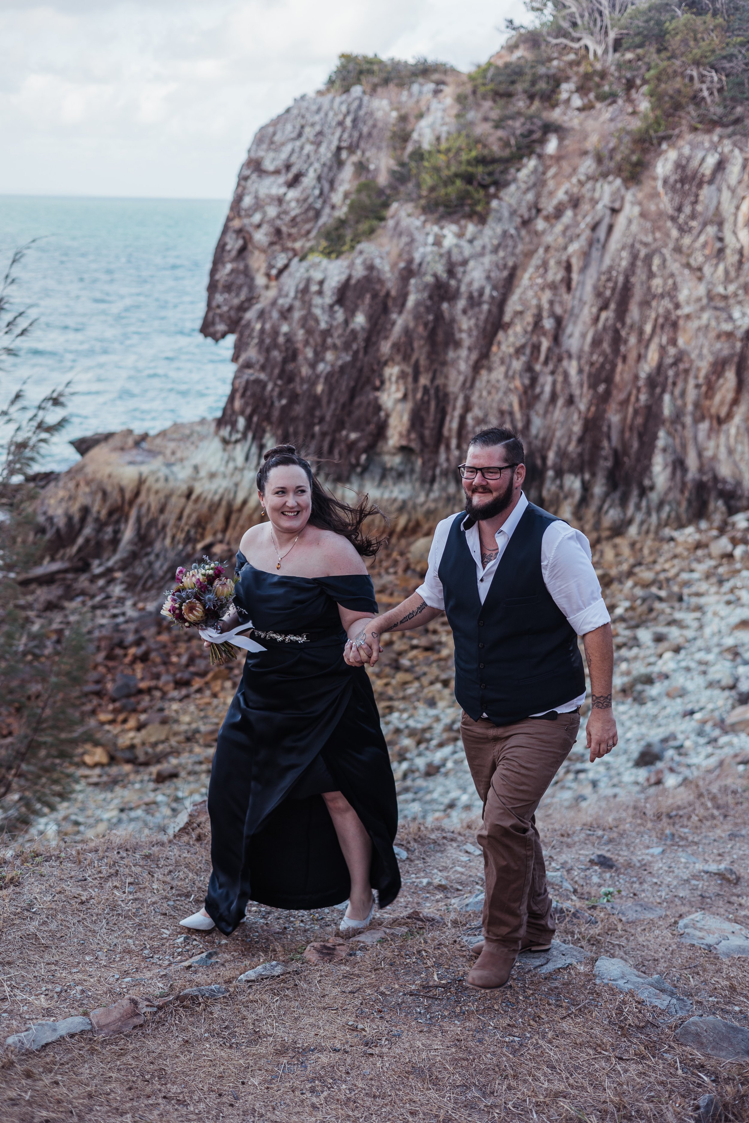 A smiling woman wearing a black off-shoulder dress and a man in a white shirt with a black vest and glasses, holding hands and walking along a rocky path near the coast with a cliff and sea in the background.