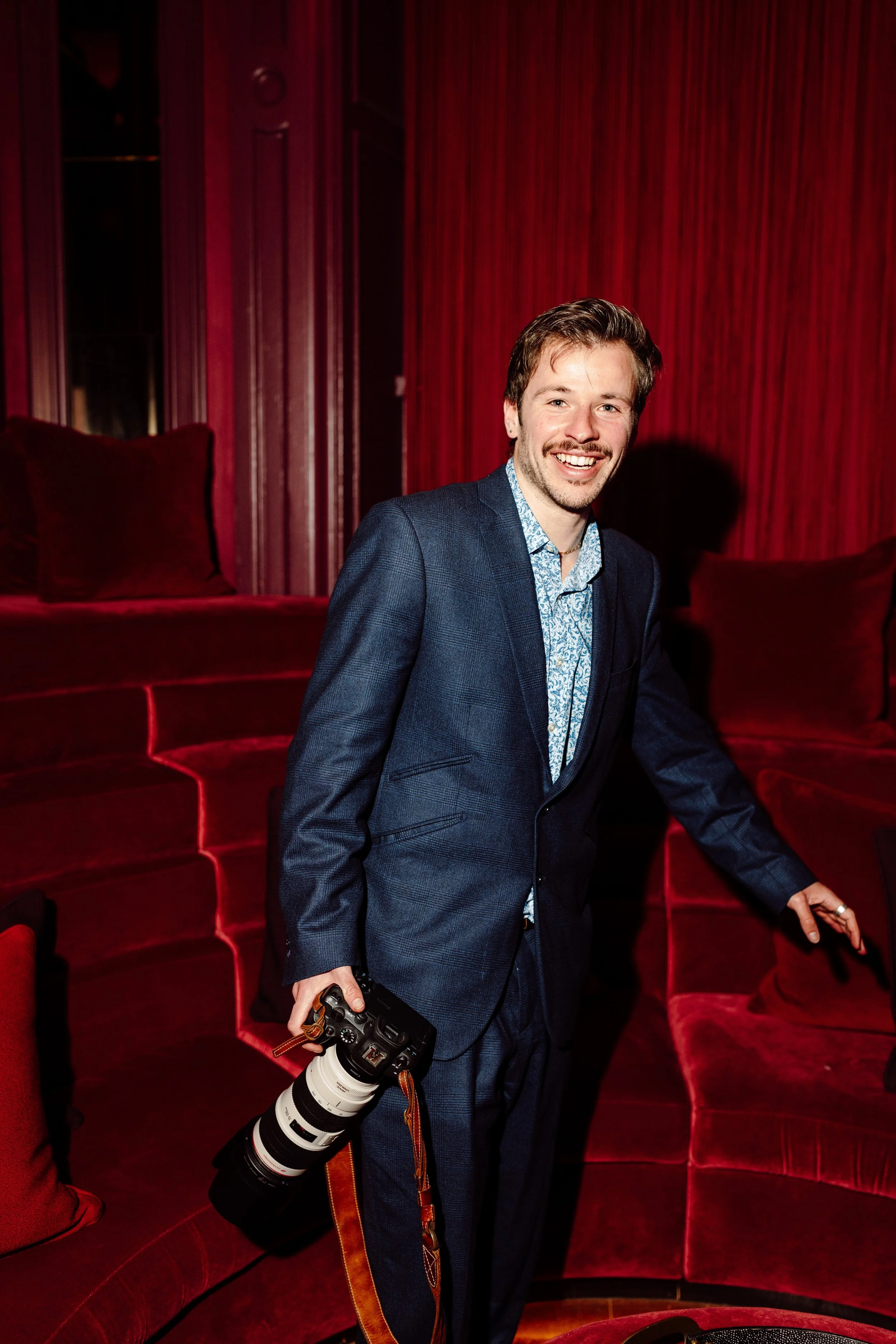 A man in a blue suit stands in front of a red velvet theater seat, holding a professional camera with a large telephoto lens, smiling at the camera, with a red velvet curtain background.