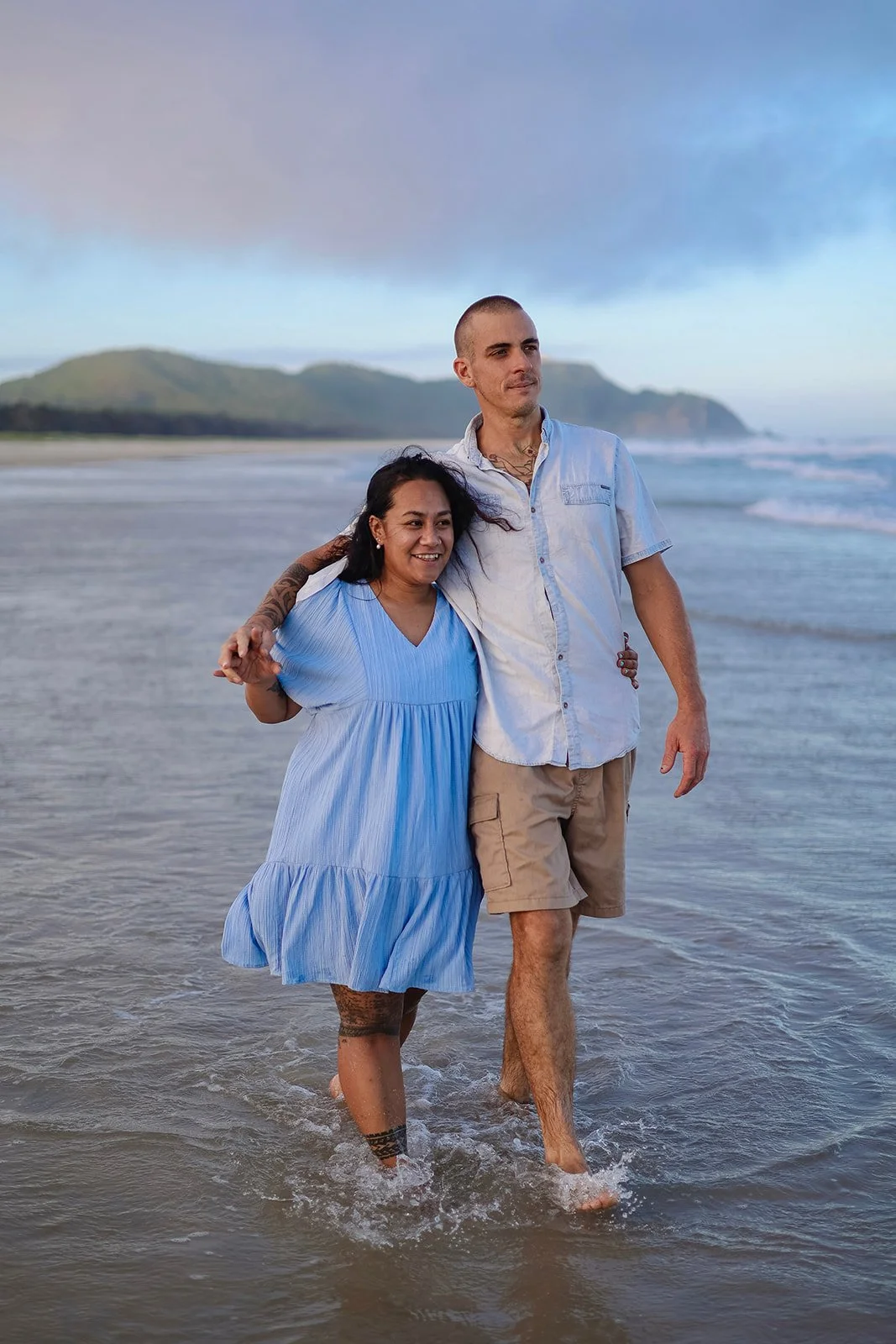 A couple walking barefoot in the ocean at sunset, with a mountainous coastline in the background, holding hands and smiling.