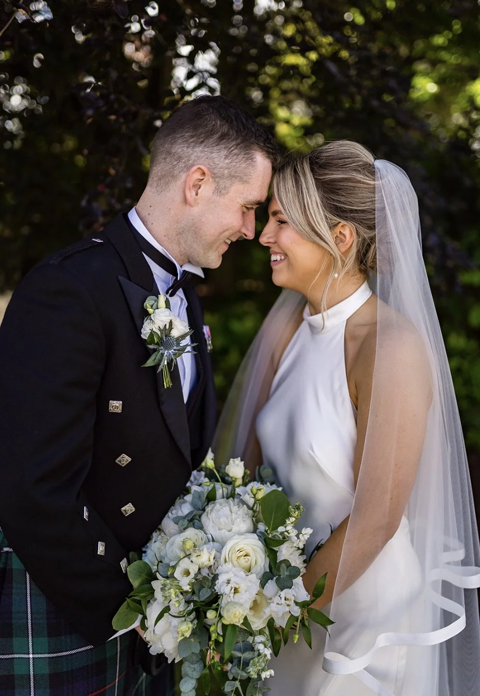 A bride and groom stand close with foreheads touching, smiling, outdoors, wedding attire, bride holding a bouquet of white flowers and greenery.