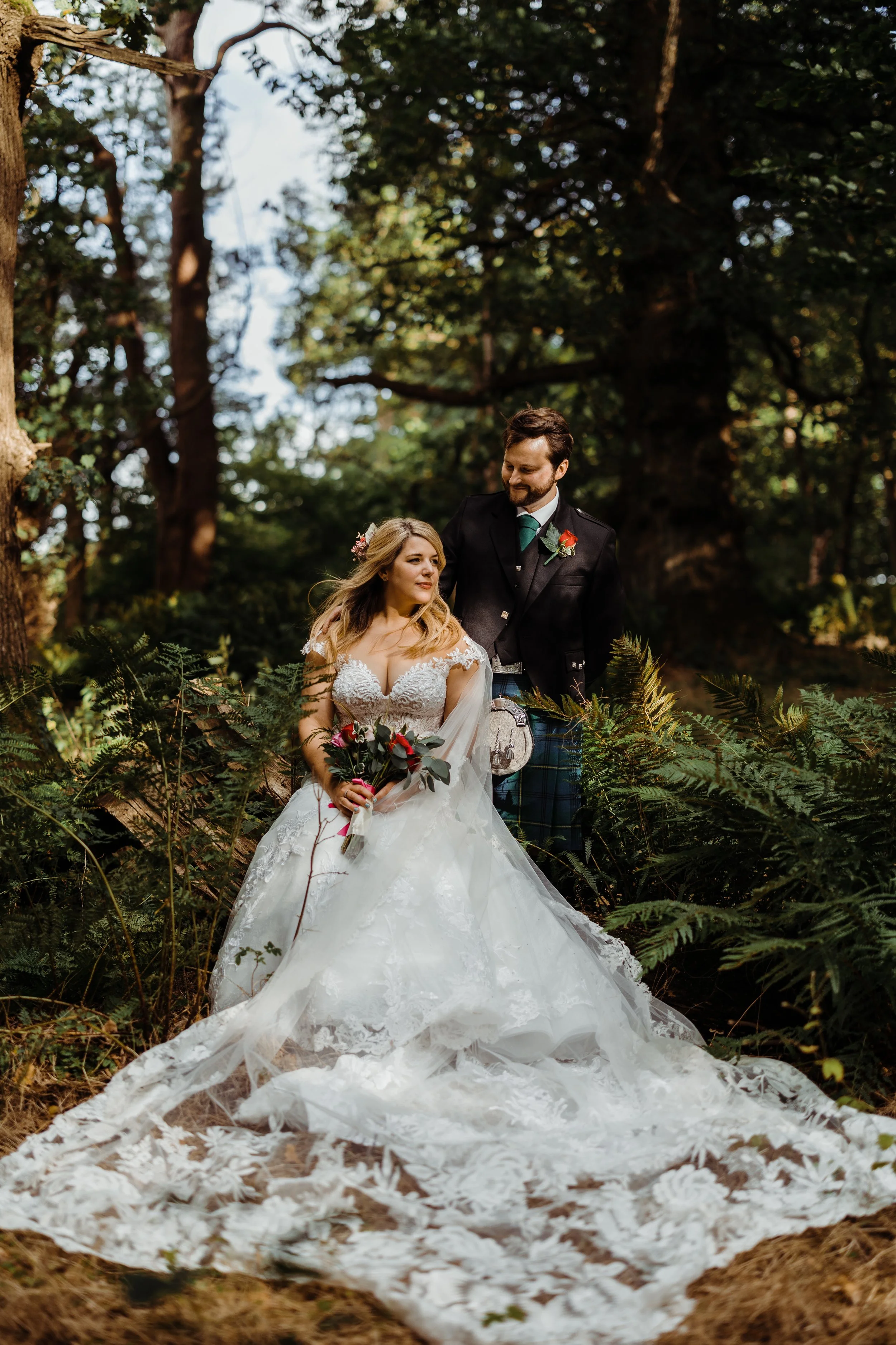 A bride and groom in wedding attire standing in a dense forest surrounded by green ferns and trees, with the bride holding a bouquet of flowers.