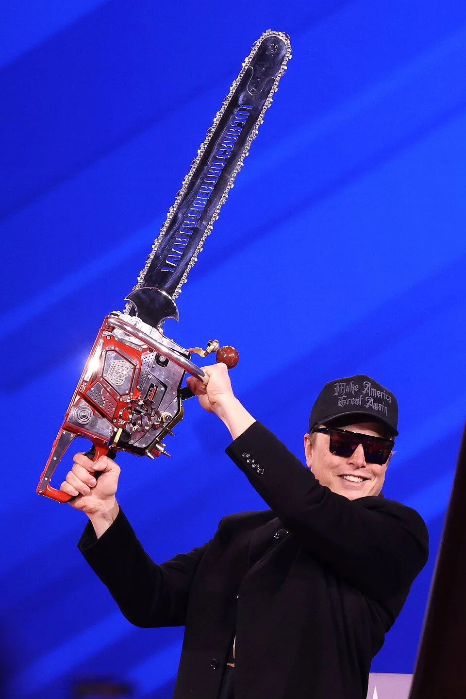 A crazed Elon Musk wearing sunglasses and a black cap with embroidered text, holding a large, modified chainsaw weapon with a metal body, against a plain blue background.