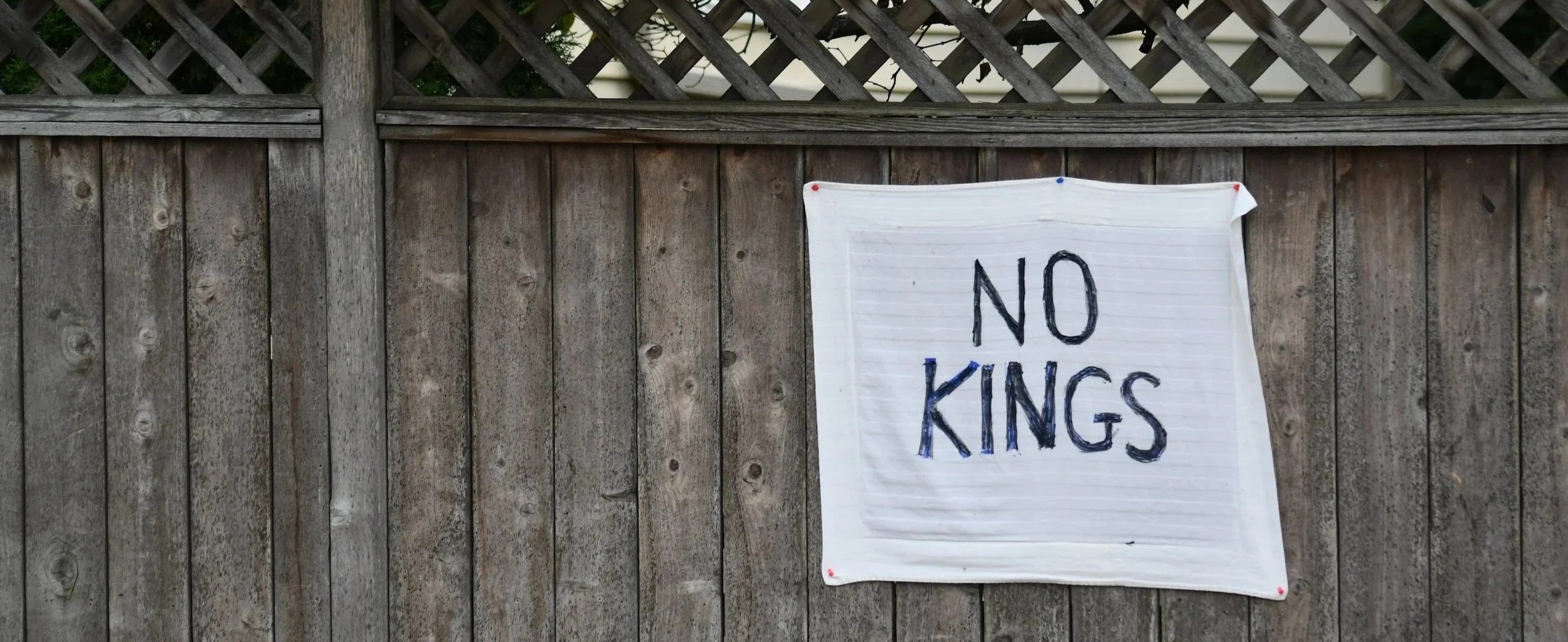 A white cloth sign with black handwritten letters saying 'NO KINGS' is attached to a wooden fence.