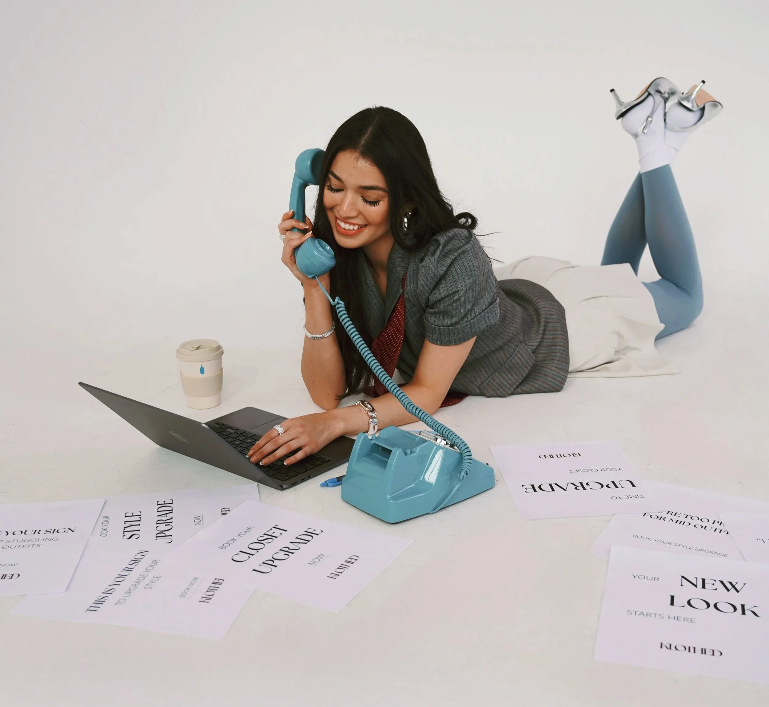 A woman lying on her stomach on a white table, talking on a blue vintage rotary phone while looking at her laptop. She is surrounded by printed sheets of paper with text about upgrading style and logo creation. She is dressed in blue tights, a stripe