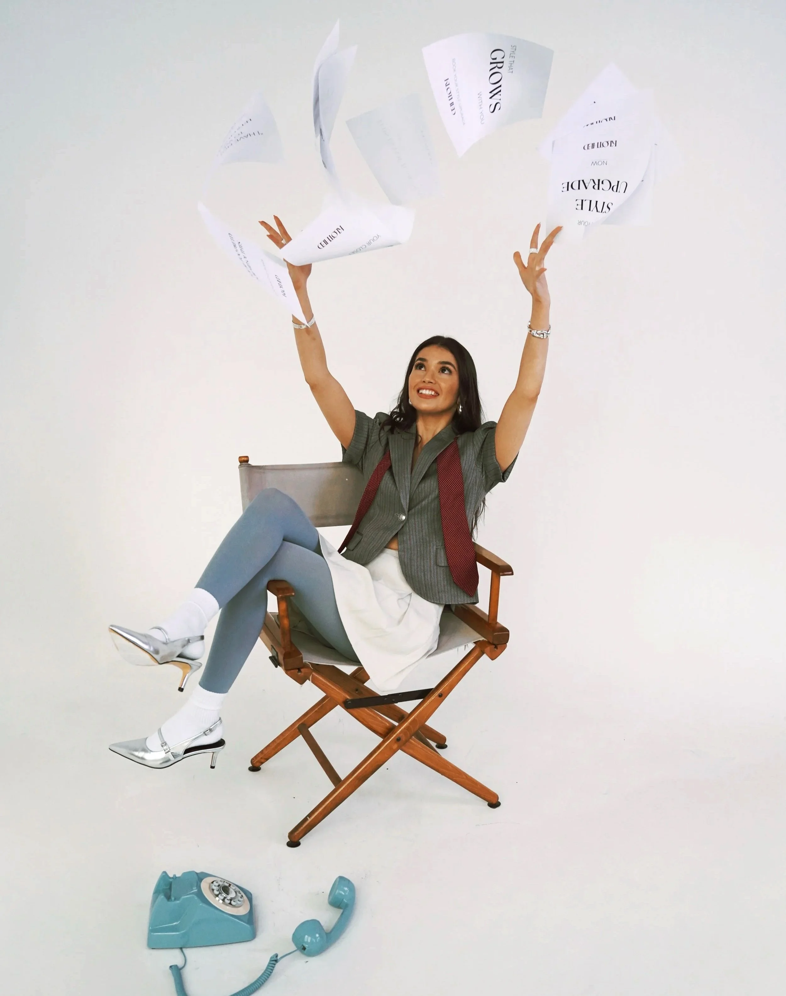A woman sitting on a wooden director's chair with her feet propped up, smiling and throwing papers into the air, with a broken vintage teal rotary telephone on the floor in front of her against a plain white background.