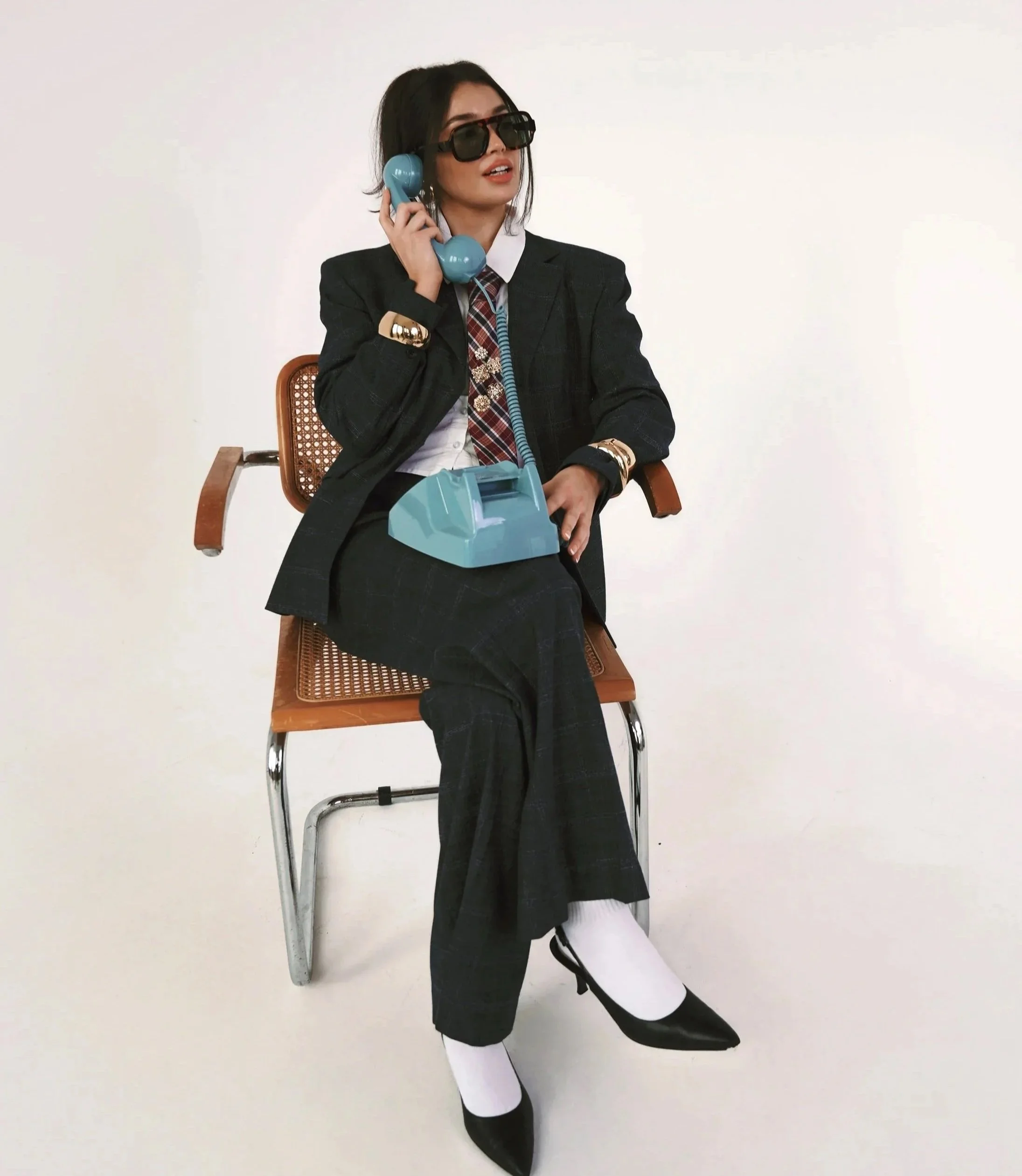 Woman in a black blazer, white shirt, plaid tie, black skirt, white tights, and black shoes sitting on a chair against a plain white wall, talking on a blue rotary phone and wearing large sunglasses.
