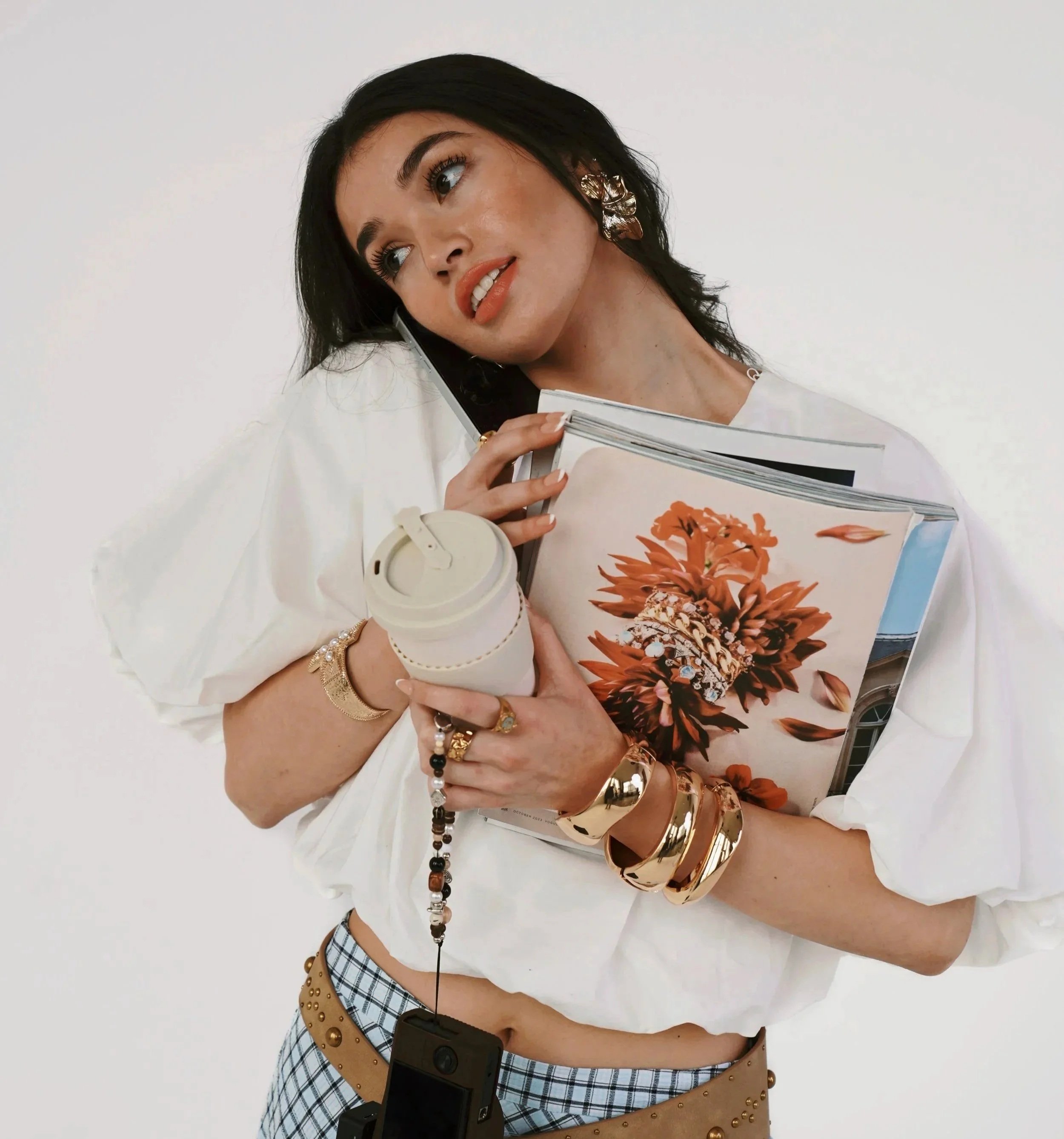 A woman with dark hair and earrings is holding a large open book with floral art on the pages, wearing a white top and plaid shorts, and carrying a beige studded bag. She is standing against a plain background.