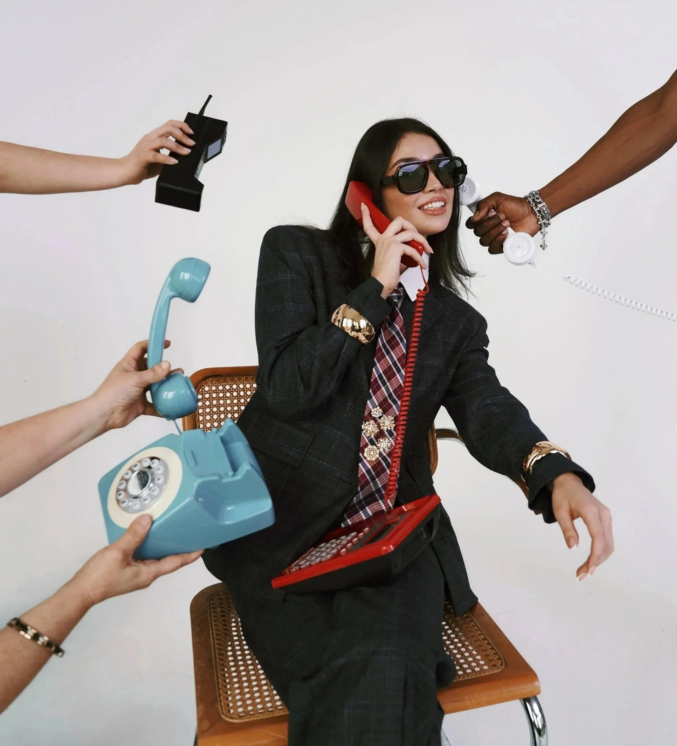 Woman with black hair, wearing sunglasses and a dark plaid blazer, talking on a red telephone, sitting on a wooden chair with a cane design on the backrest. Multiple hands around her holding various telephones and devices.