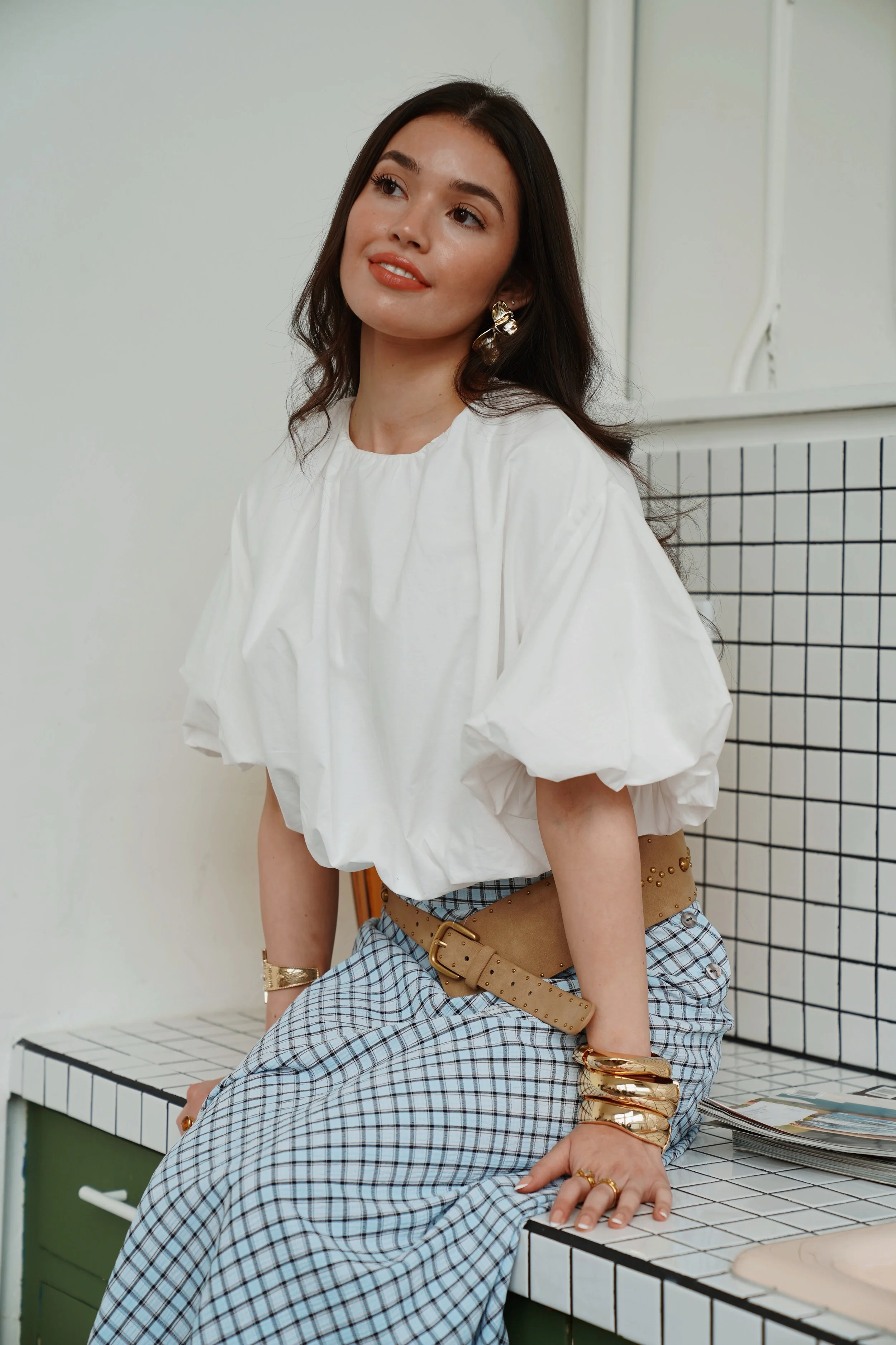 Woman with long dark hair wearing a white puff-sleeve blouse and plaid wide-leg pants, sitting on a tiled counter with fashion jewelry, looking away, in a minimalist room.