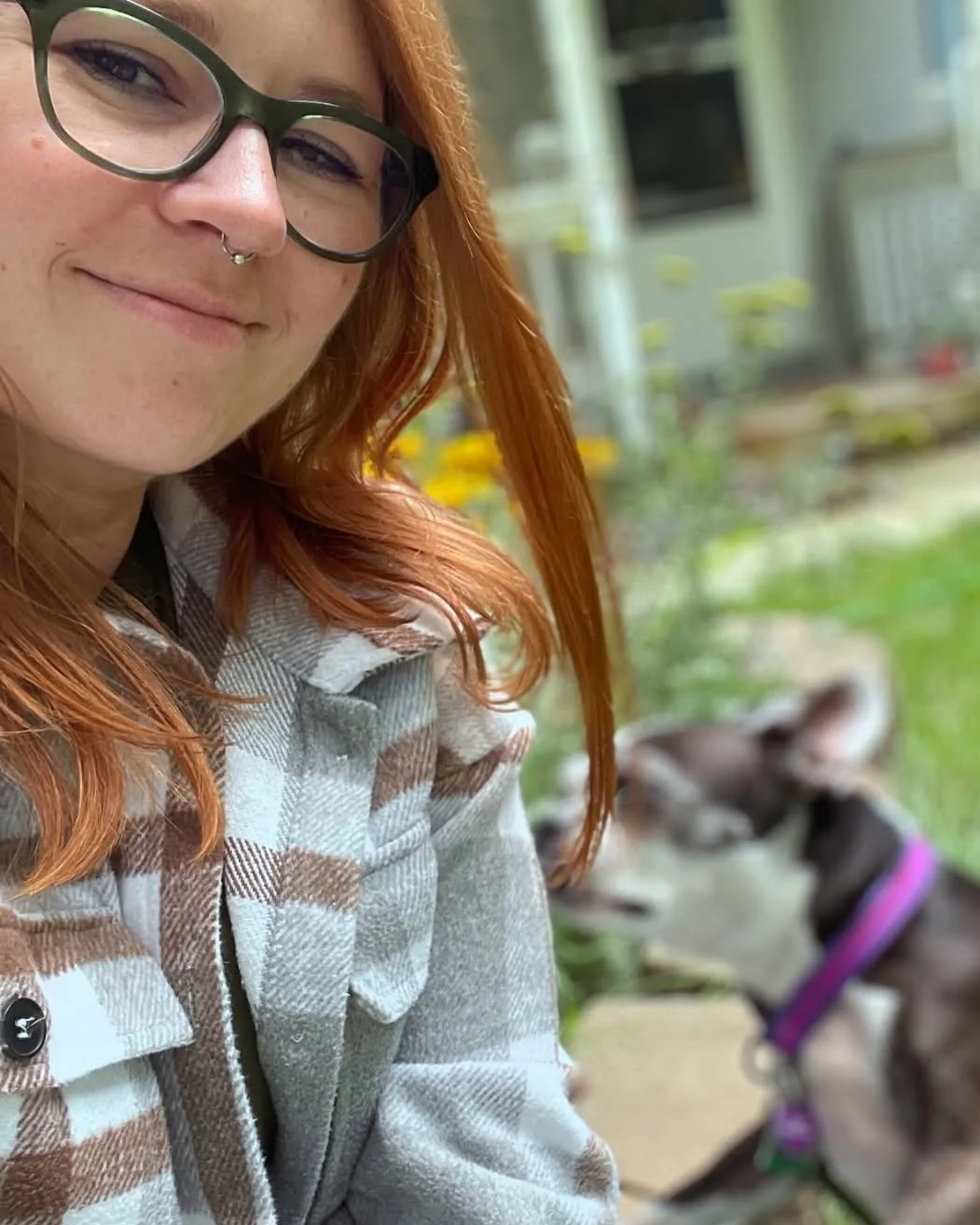 A woman with glasses and a septum piercing taking a selfie outdoors, with a dog in the background.