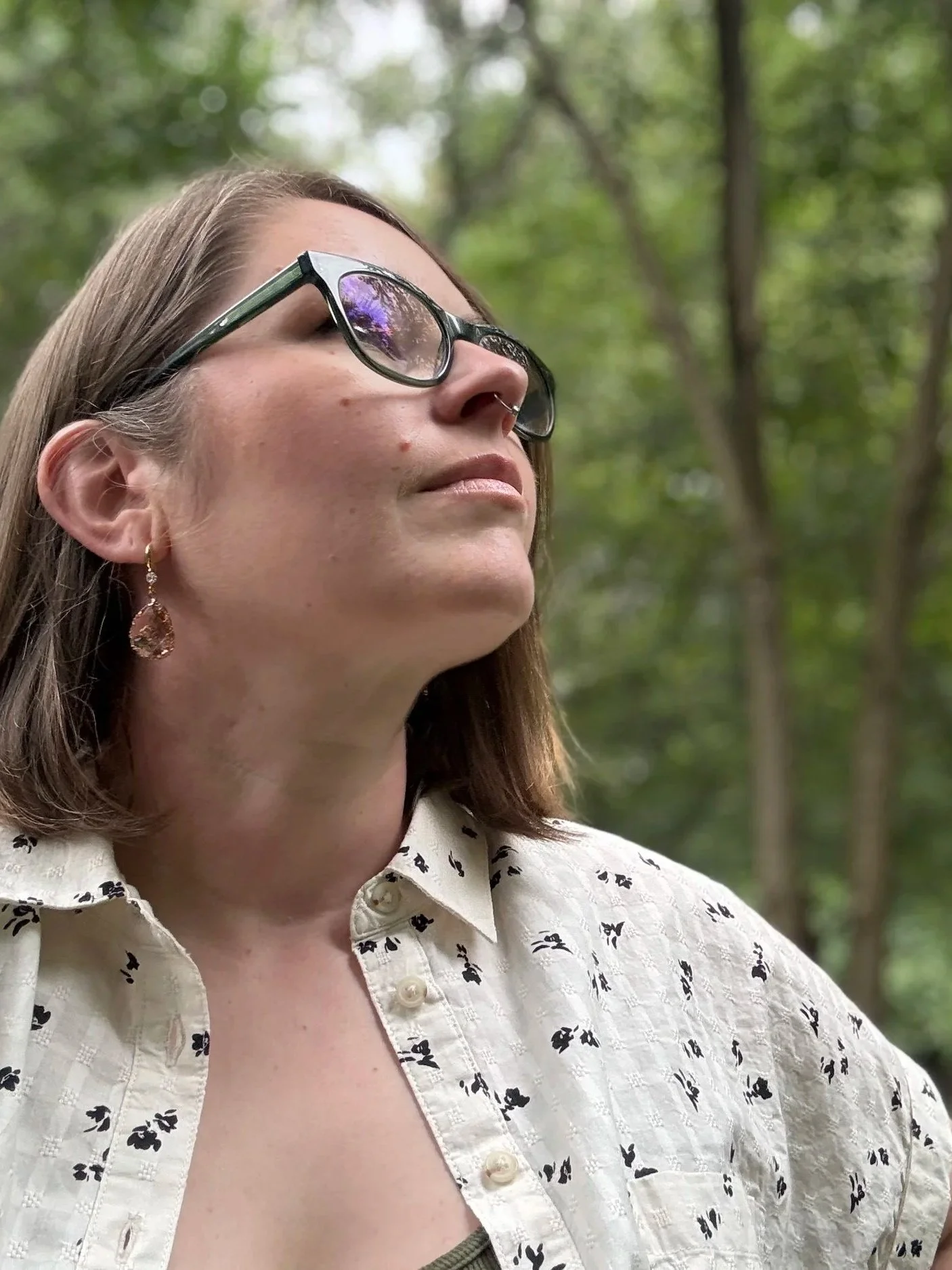 A woman with brown hair wearing glasses and gold earrings, looking up outdoors surrounded by green trees, dressed in a cream-colored shirt with black floral pattern.