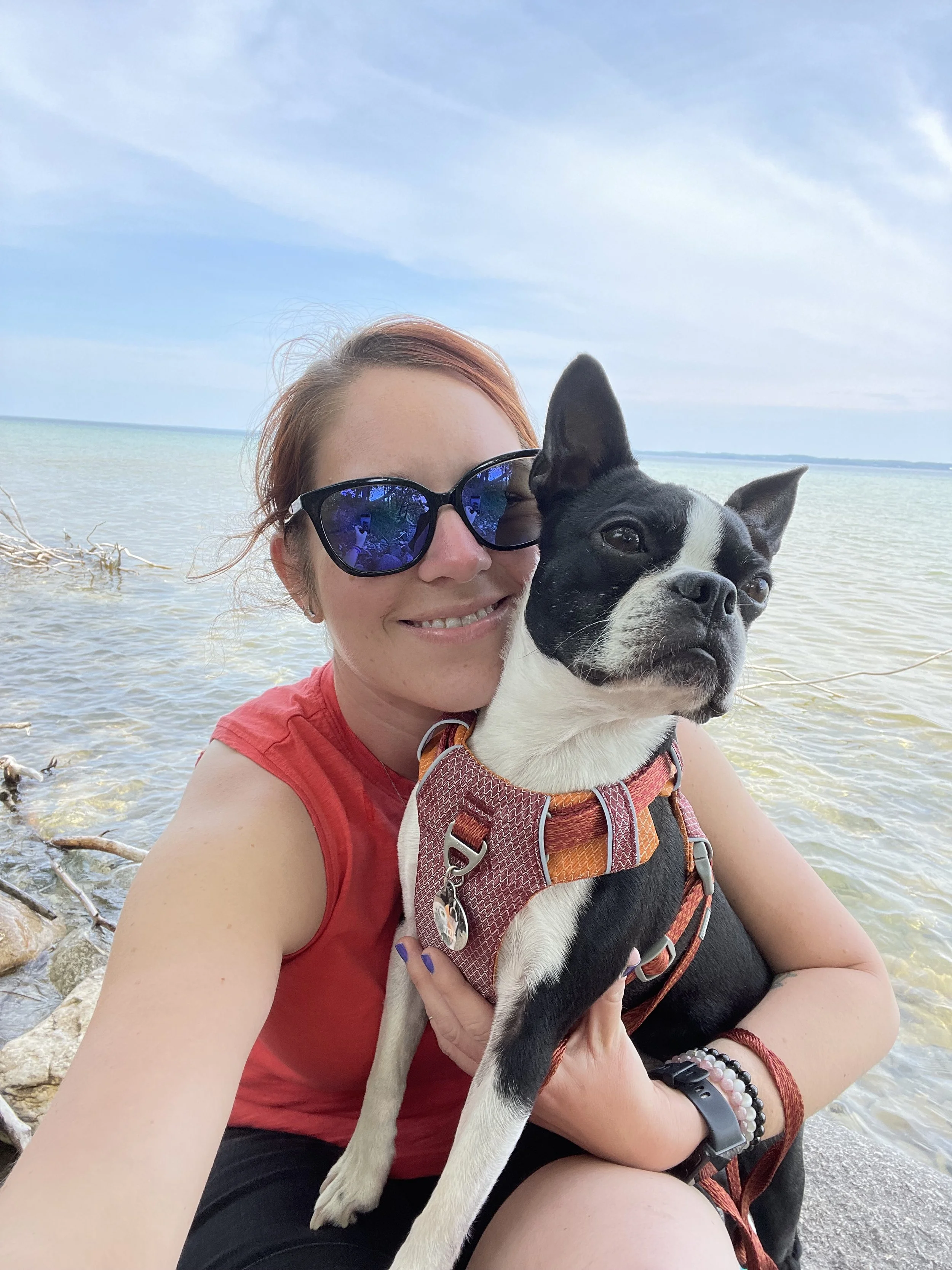 A woman with red hair wearing sunglasses and a red tank top, holding a black and white Boston Terrier dog at the beach with water and a blue sky in the background.