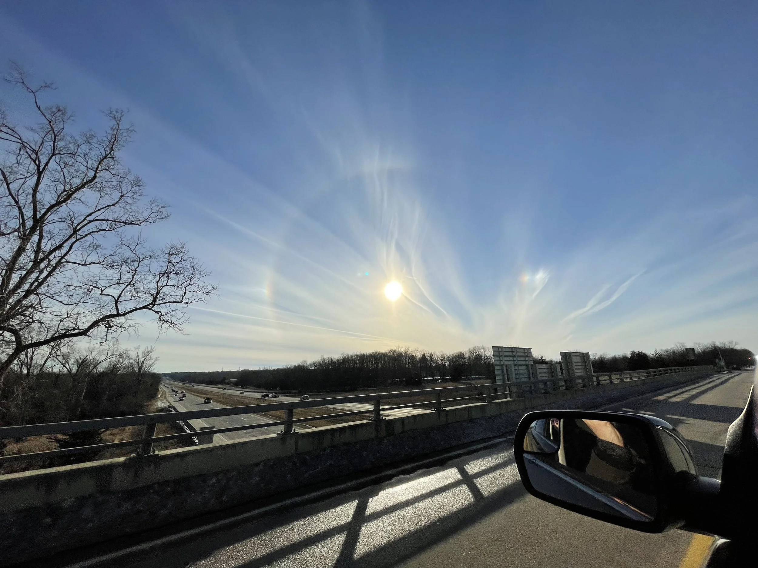 View of a sun dog in a bright blue sky and a highway with multiple lanes filled with cars, seen from the passenger side of a vehicle. Trees without leaves and buildings are visible in the distance. The sky is clear with the sun shining brightly.