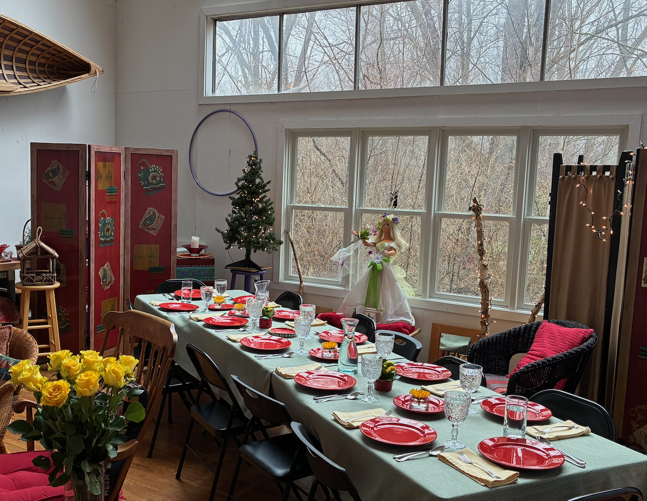 A dining room prepared for a holiday celebration with a long table set with red plates, glassware, and napkins. A small decorated Christmas tree, a doll figure, and seasonal decorations are visible near large windows showing a wintery outdoor scene.