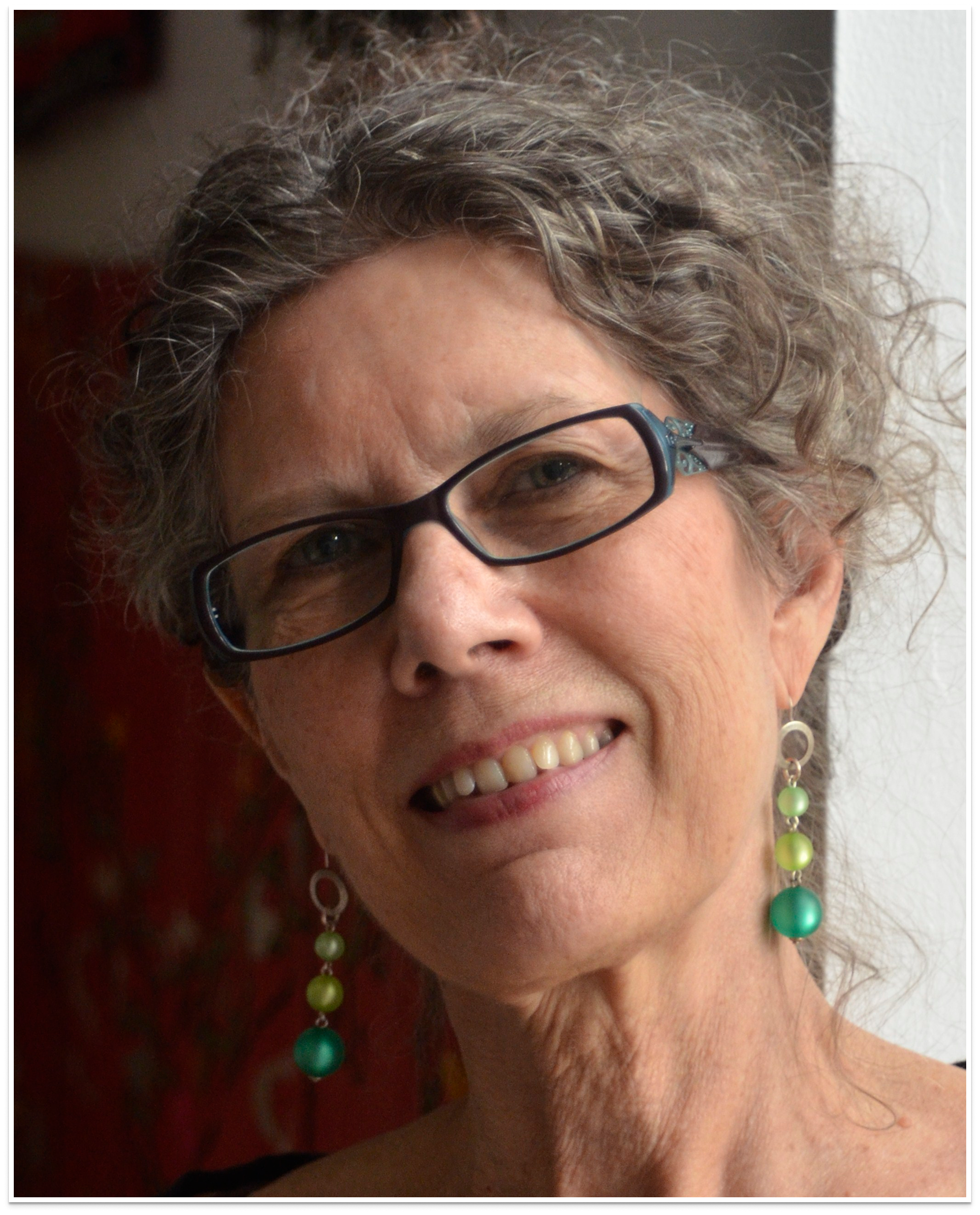 Close-up of a woman with curly gray hair and glasses, smiling, wearing colorful bead earrings.