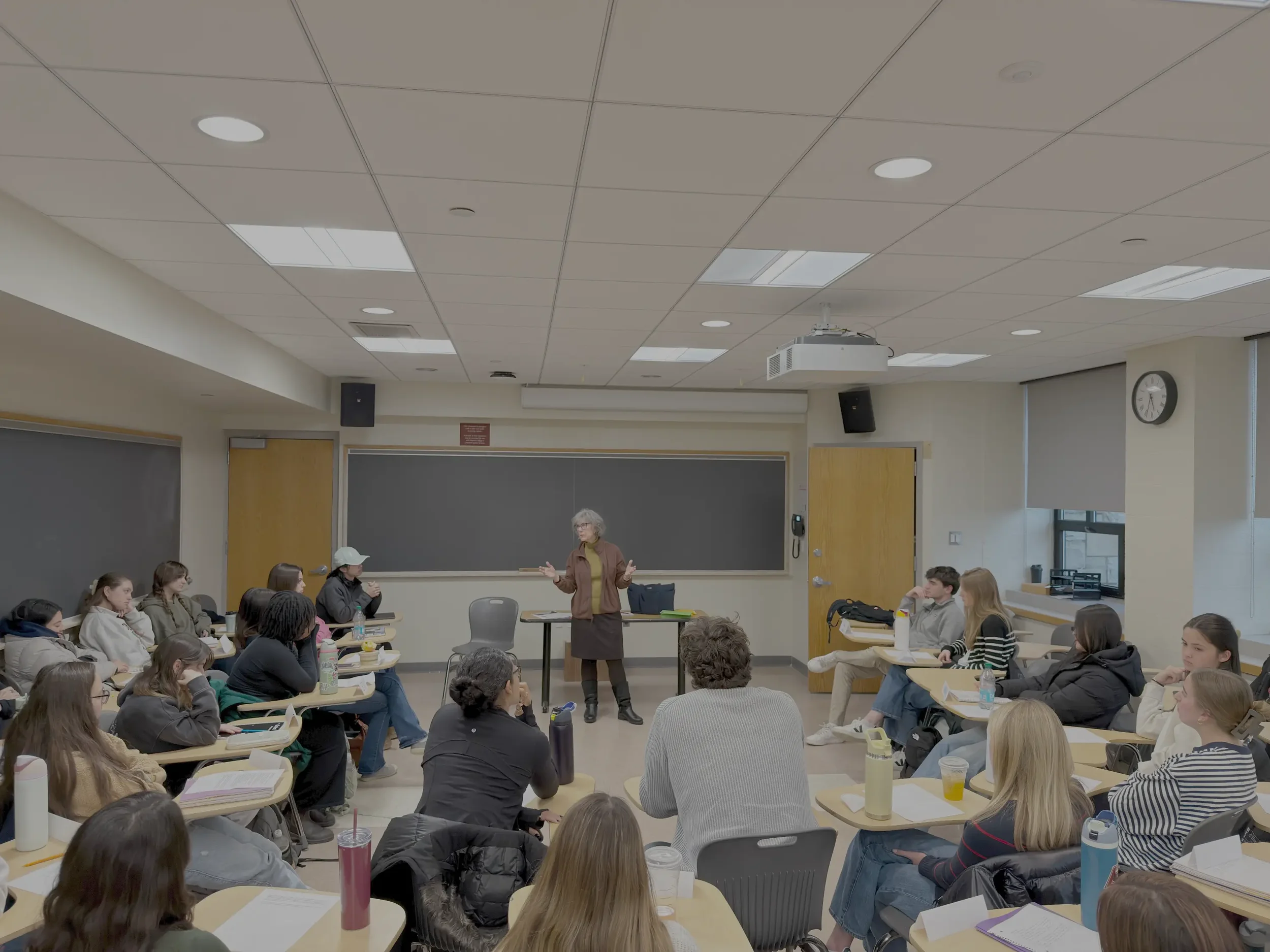 An instructor or speaker standing at the front of a classroom full of students, gesturing with her hands. The classroom has a ceiling with lights and a projector, and the students are seated at tables, listening attentively.