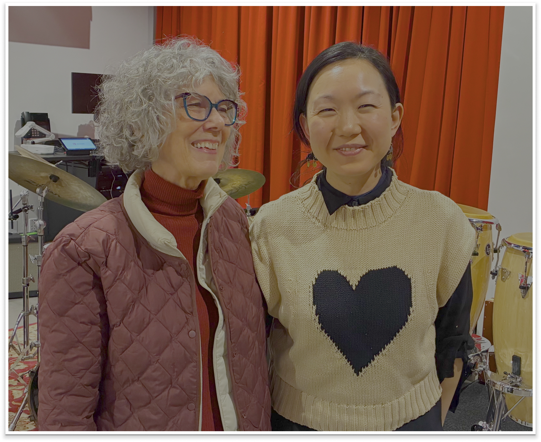 Two women posing together in a room with musical instruments, including drums, in the background.