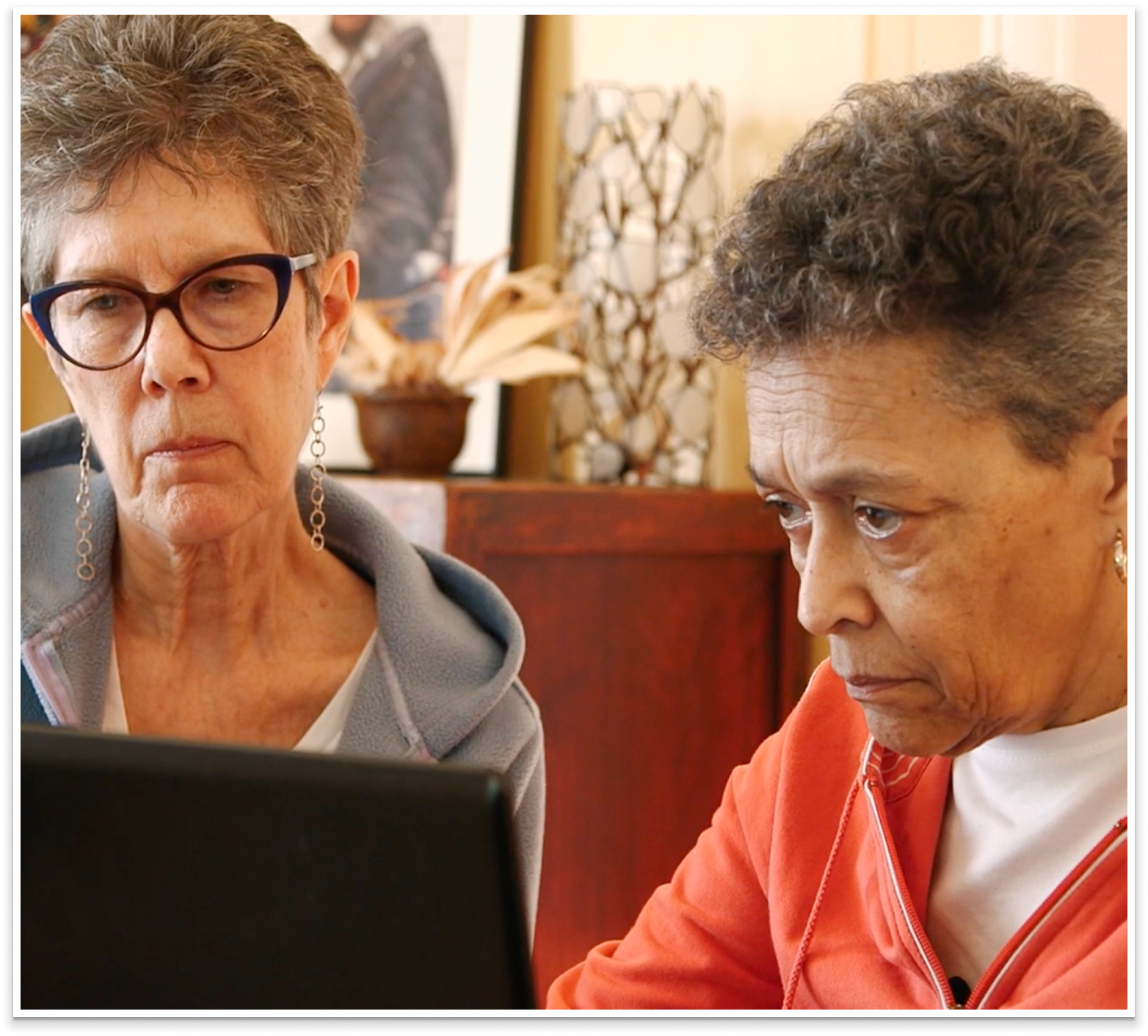 Two women are looking at a laptop screen together. One woman has short, curly hair and is wearing an orange zip-up jacket, while the other woman has short, straight hair, glasses, and is wearing a gray hoodie with a necklace.