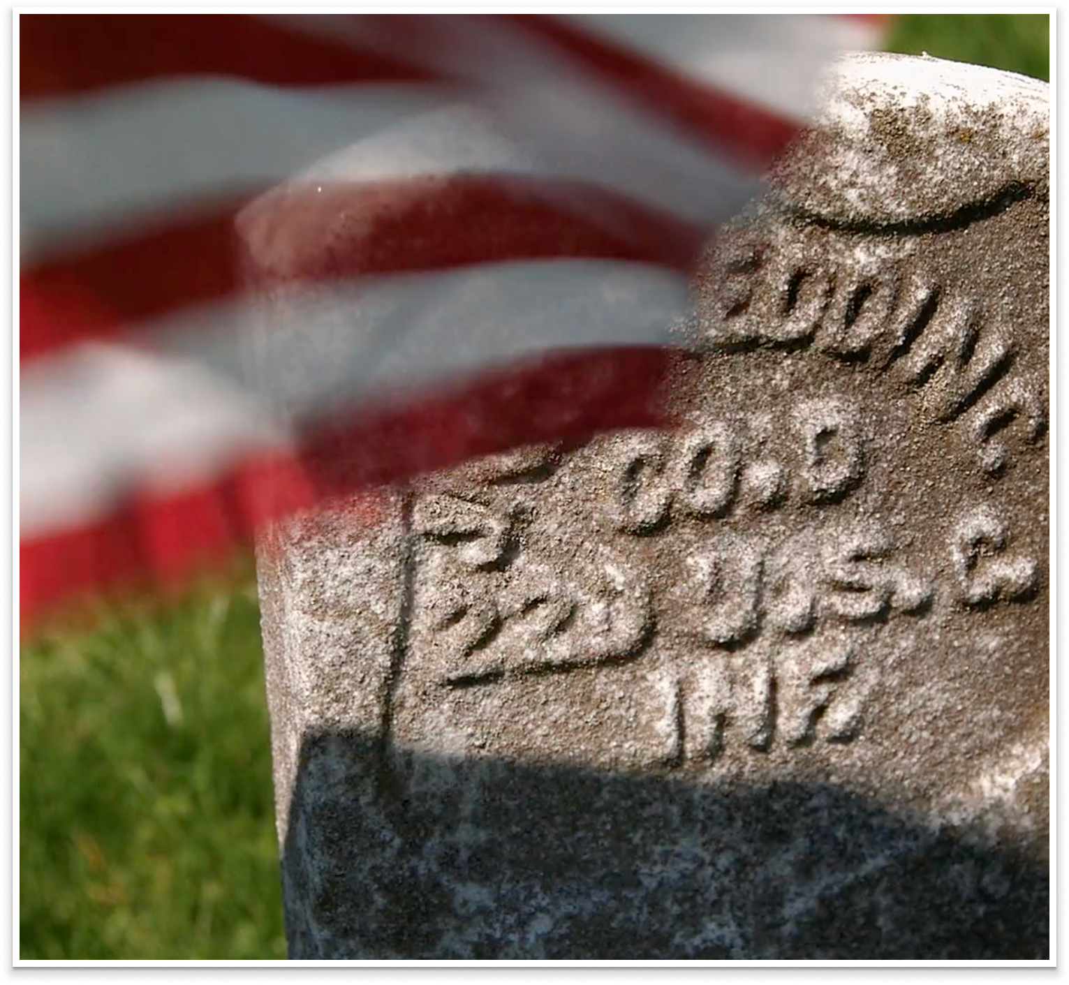 Close-up of a weathered stone memorial or plaque with engraved text, partially obscured by an American flag. The flag is draped over or near the stone, with red and white stripes visible.