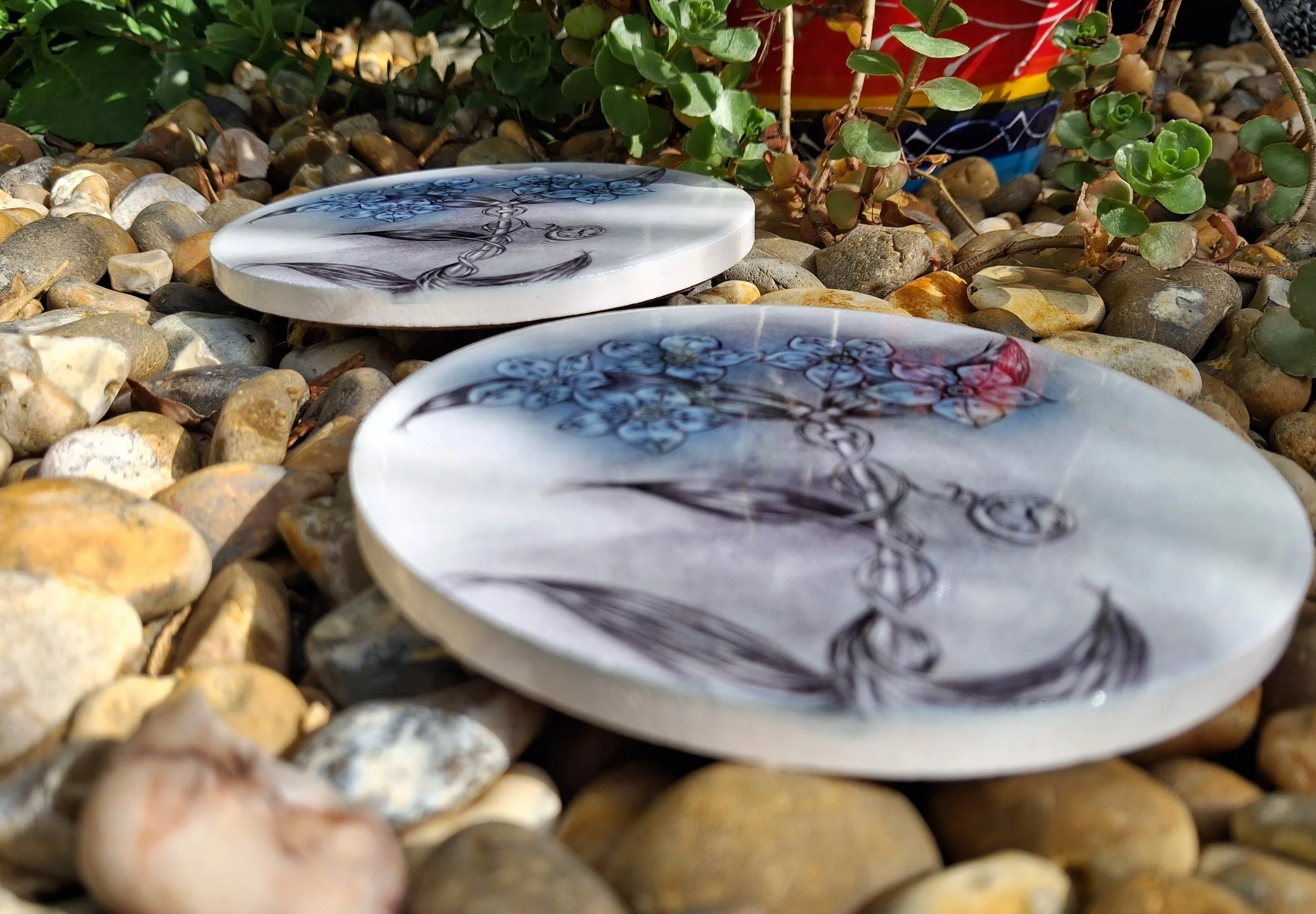 two round ceramic coasters placed on pebbles in nice garden area, the white coasters have a hand drawn forget me not flower printed into them, with blue petals